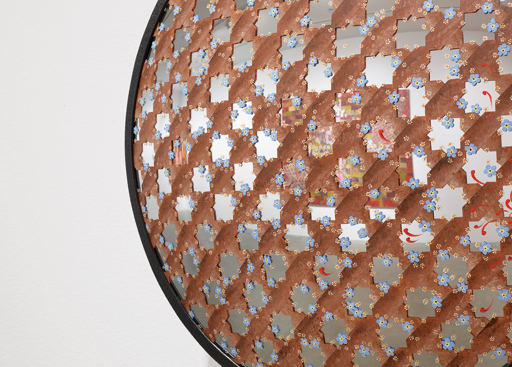 A close-up detail of a round convex security mirror that is covered by an interlocking brown painted star pattern. The uncovered “stars” are the exposed bare mirror reflecting the gallery space. Dispersed amongst the star pattern are small blue and yellow flowers with red brushtrokes scattered across the mirror.