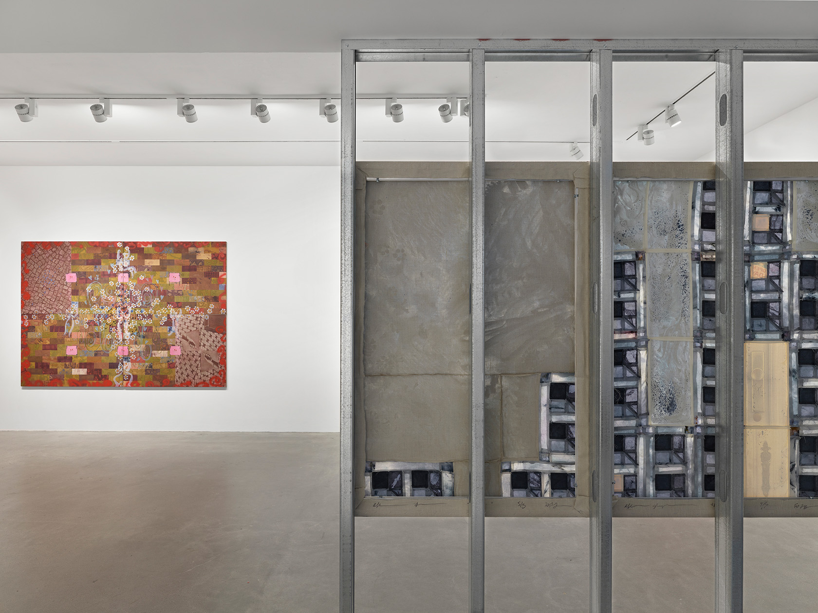 A view of a gallery space with two paintings on display. In the foreground on the right, the back side of a painting hangs across four steel beams, composed of blue patchwork pieces. In the background on the left, a painting featuring a brick pattern and flowers hangs on a white wall.