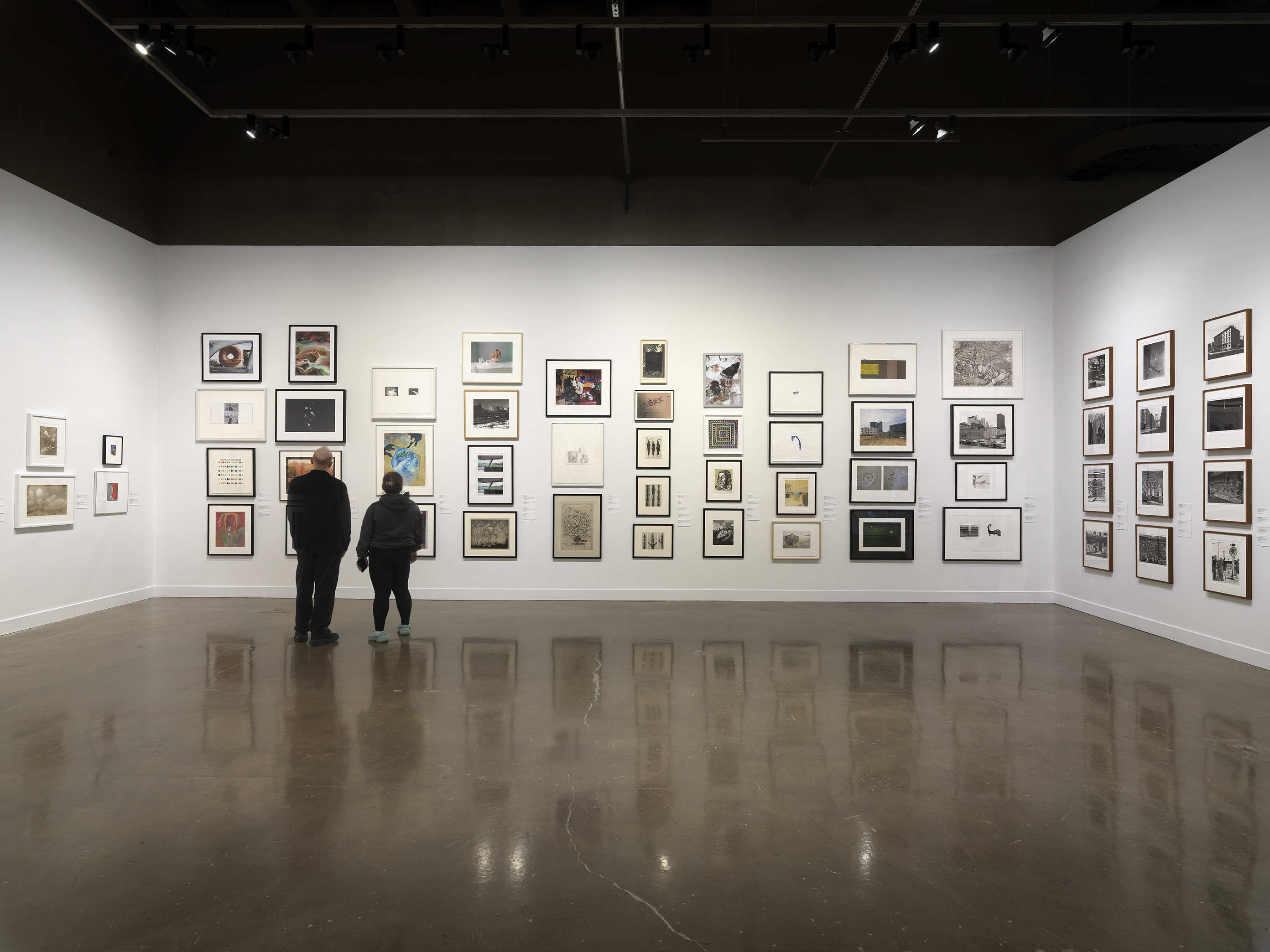 Installation view with framed artworks hanging on the walls salon style and two people standing beside each other in the center.