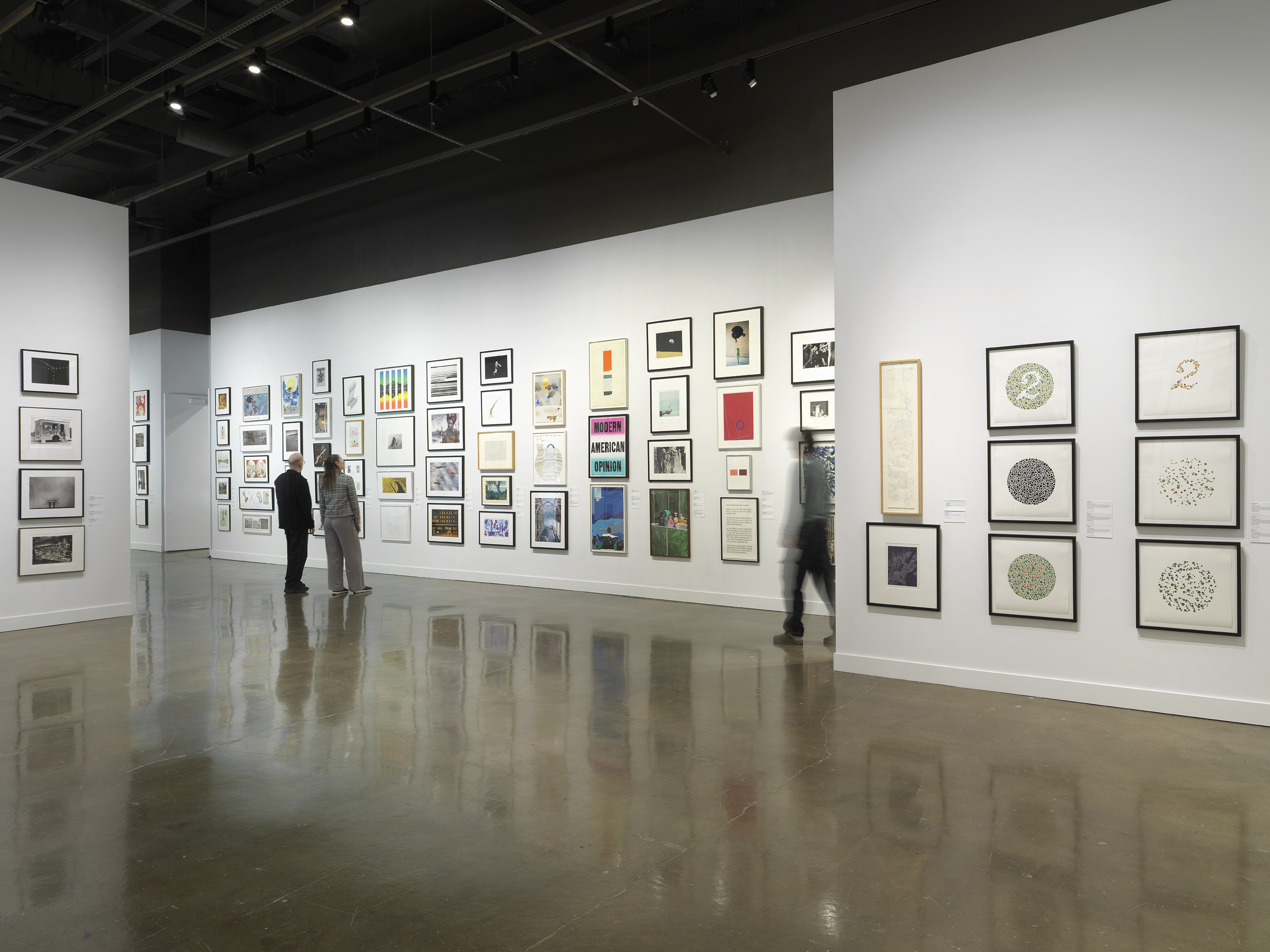 Installation view with framed artworks hanging on the walls salon style and three people standing looking at the art on the walls throughout the room.