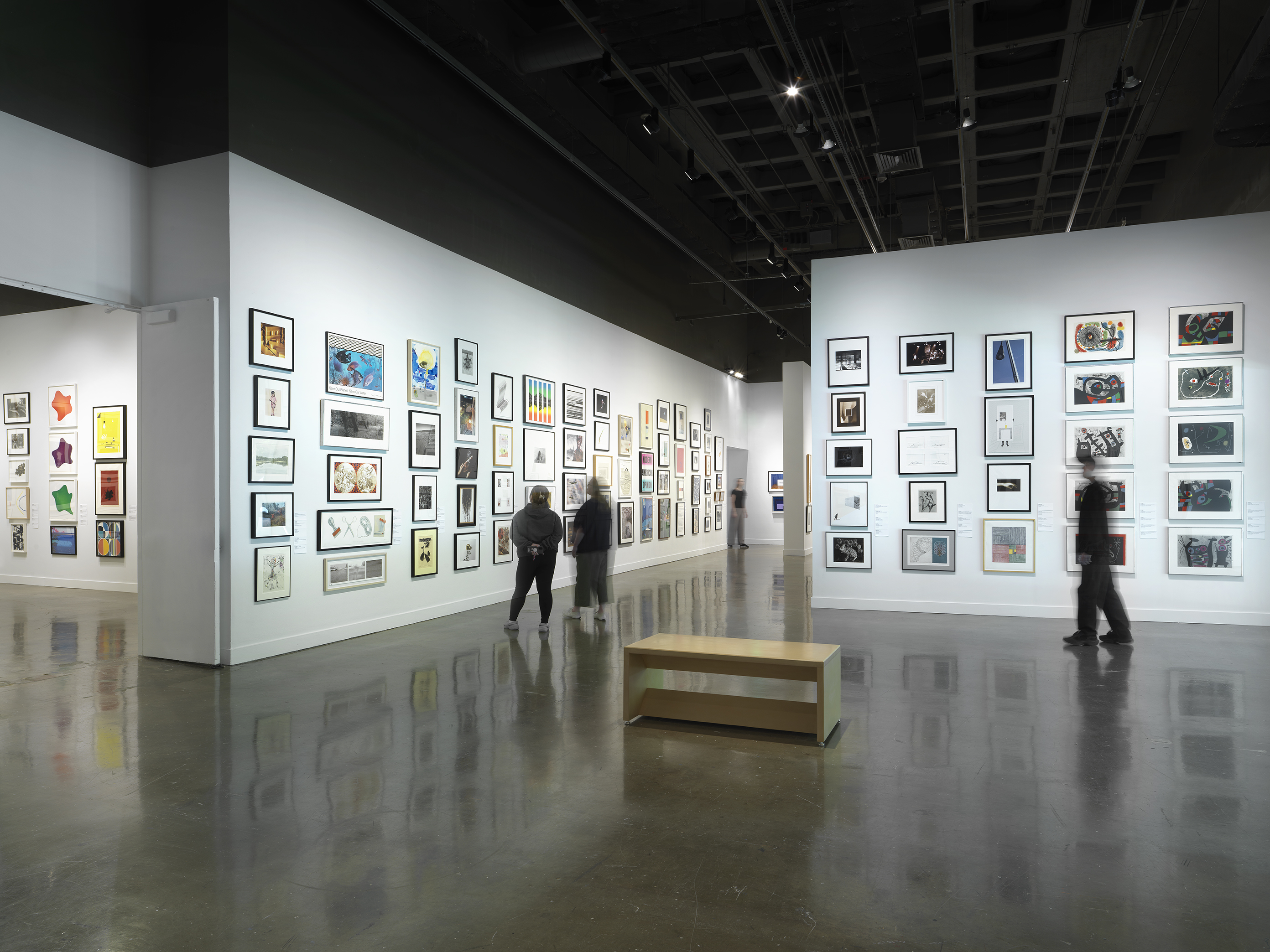 Installation view with framed artworks hanging on the walls salon style and four people standing looking at the art on the walls throughout the room.