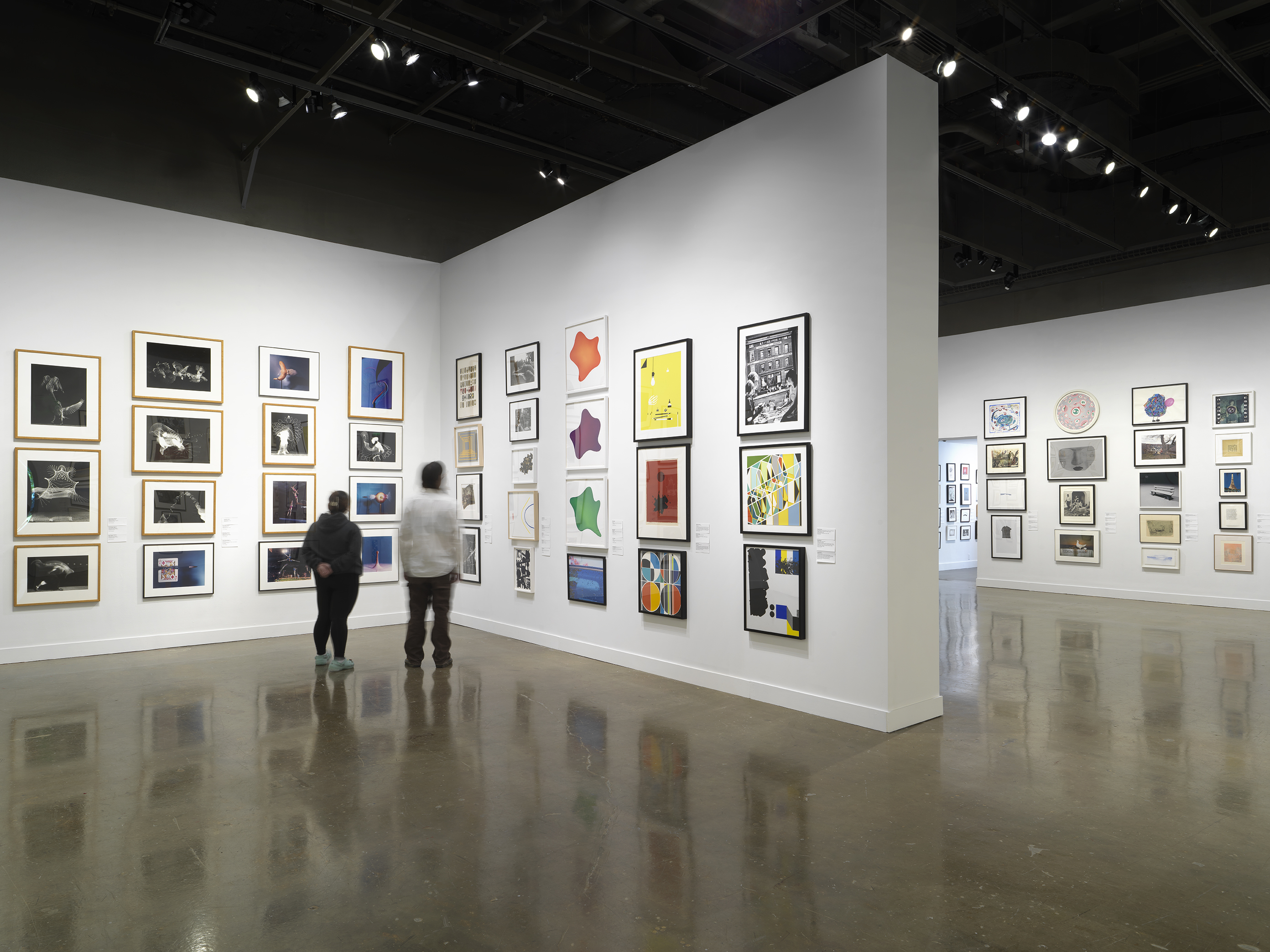 Installation view with framed artworks hanging on the walls salon style and two people standing beside each other in the center.