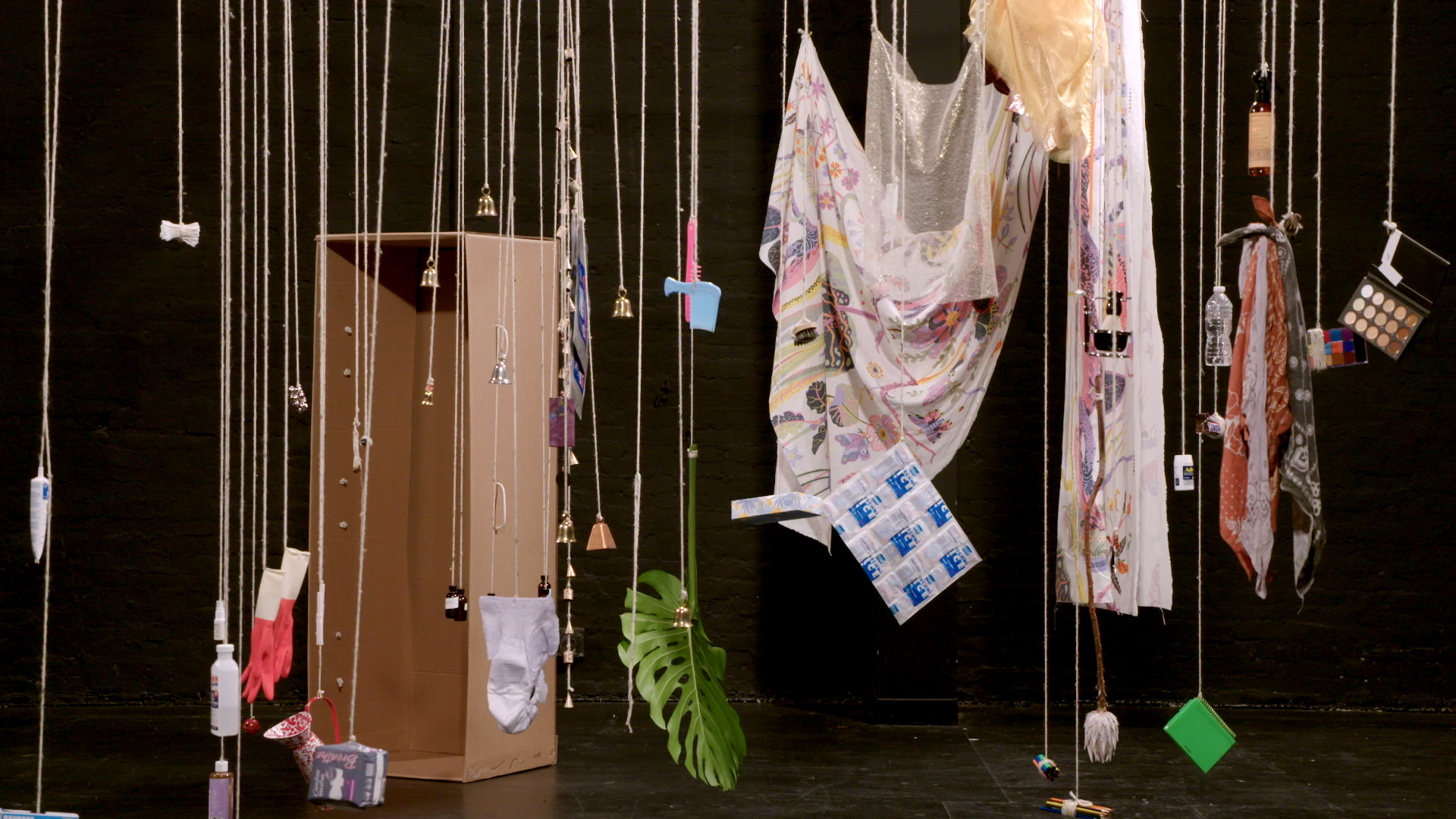 Everyday objects are suspended from the ceiling throughout the frame in front of a black background.