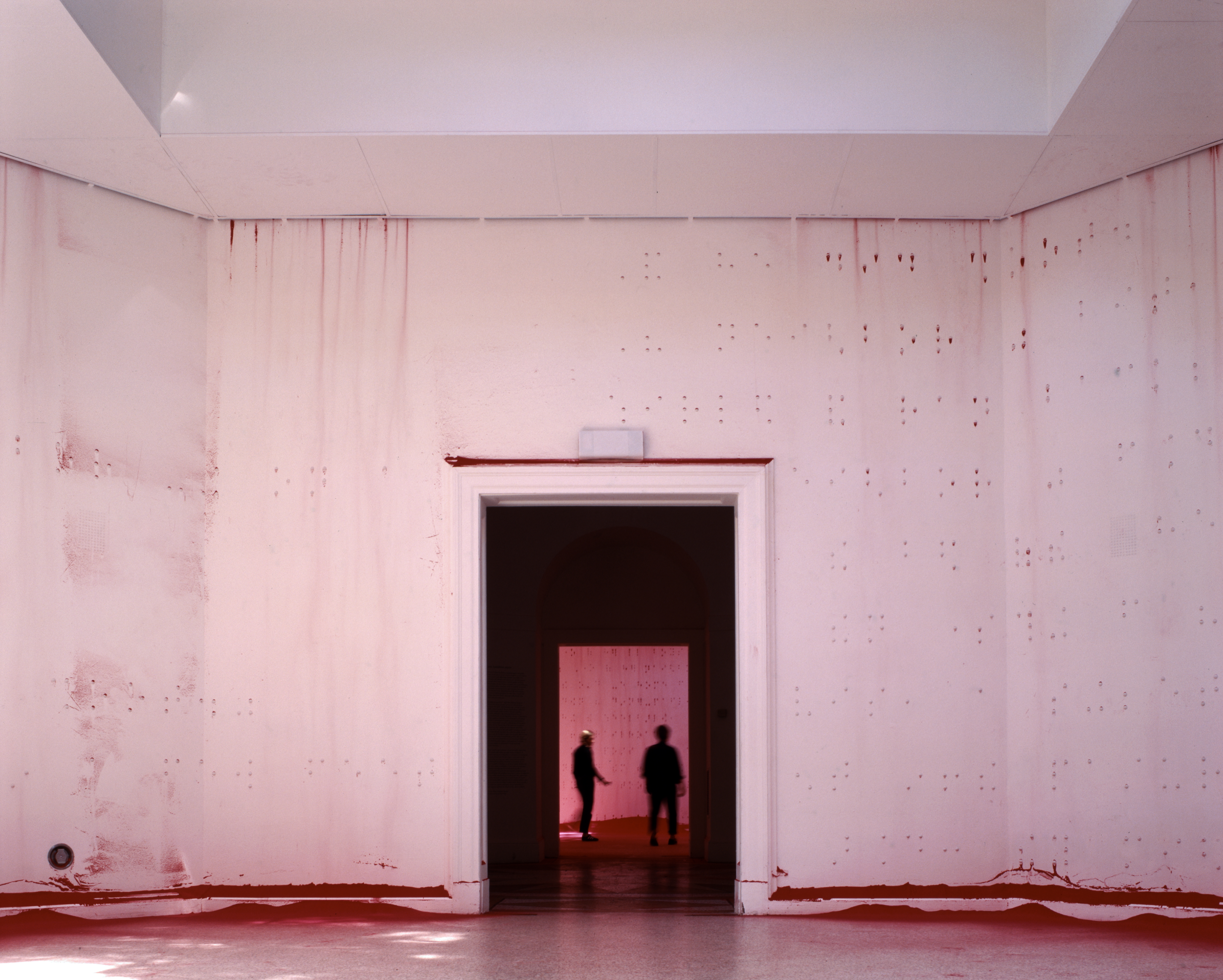An open white-walled room with braille raised bumps across the walls. The silhouettes of two people can be seen through the dark doorway in the center of the image.