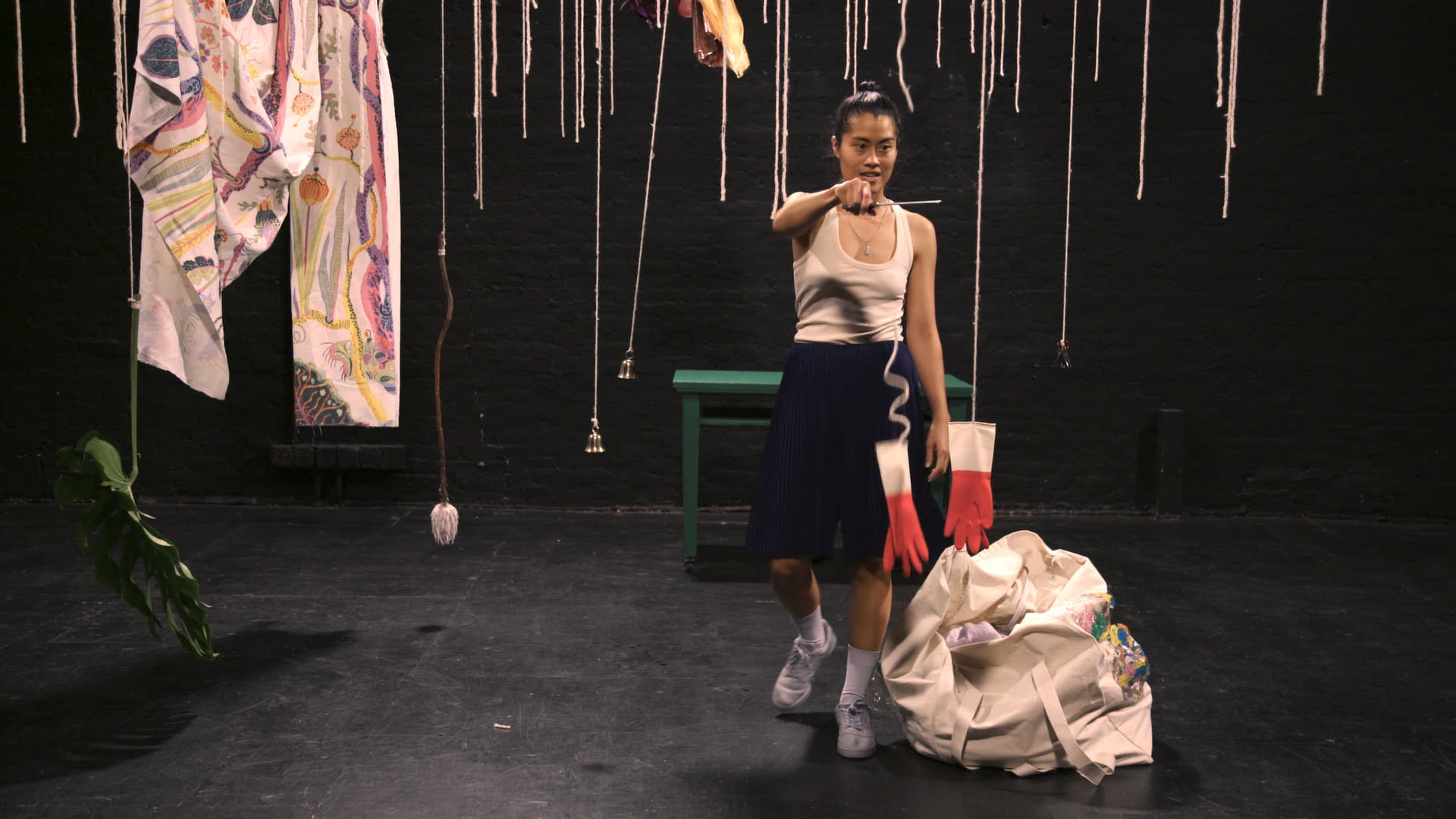 A woman is standing with scissors about to cut a string hanging from the ceiling on the right side of the frame in front of a black background. Everyday objects are suspended from the ceiling on the left side of the frame.