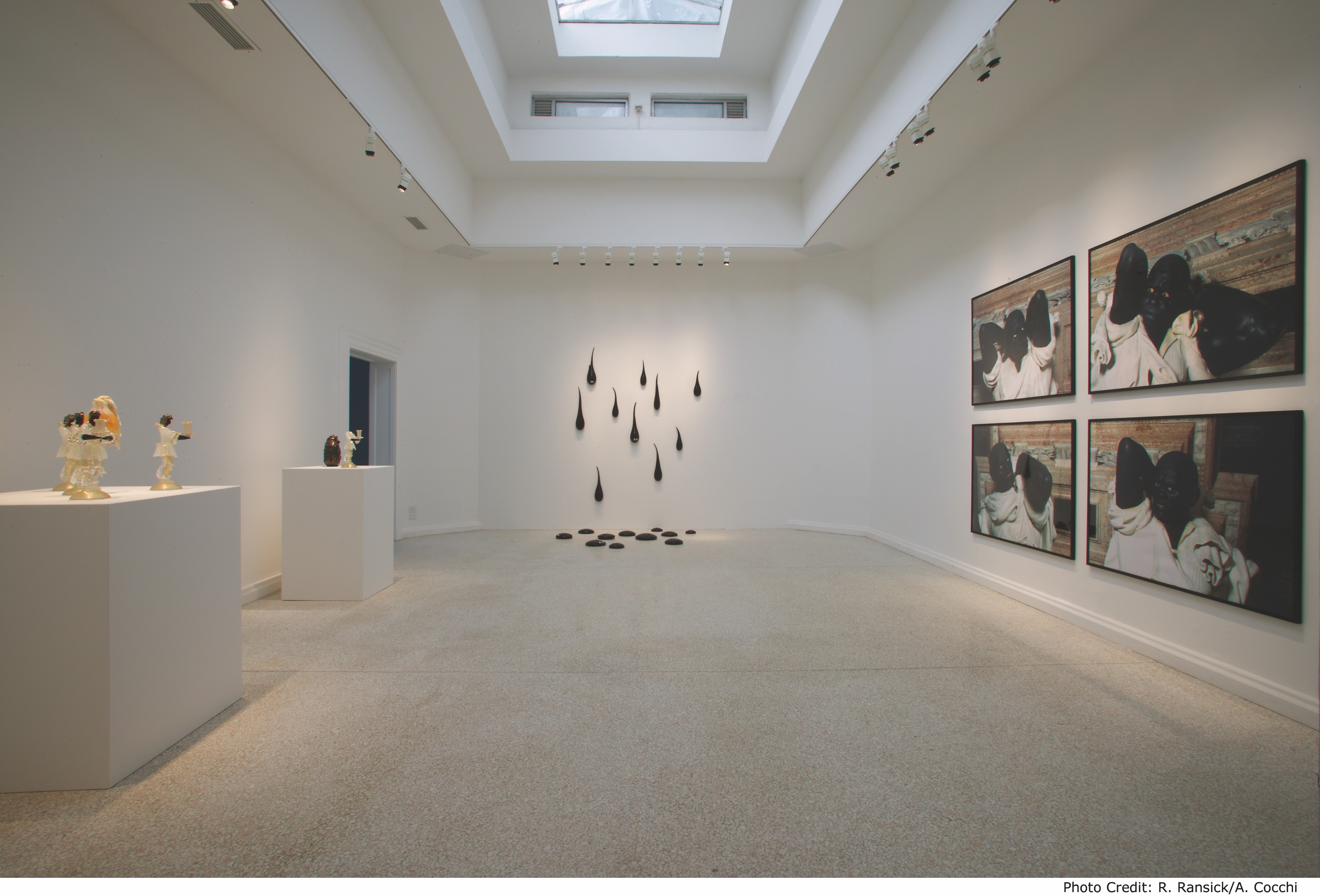 White wall room with four paintings hanging in a grid on the right side of the frame. Black oil drip sculptures are hanging in a cluster on the back wall and dripping onto the floor.