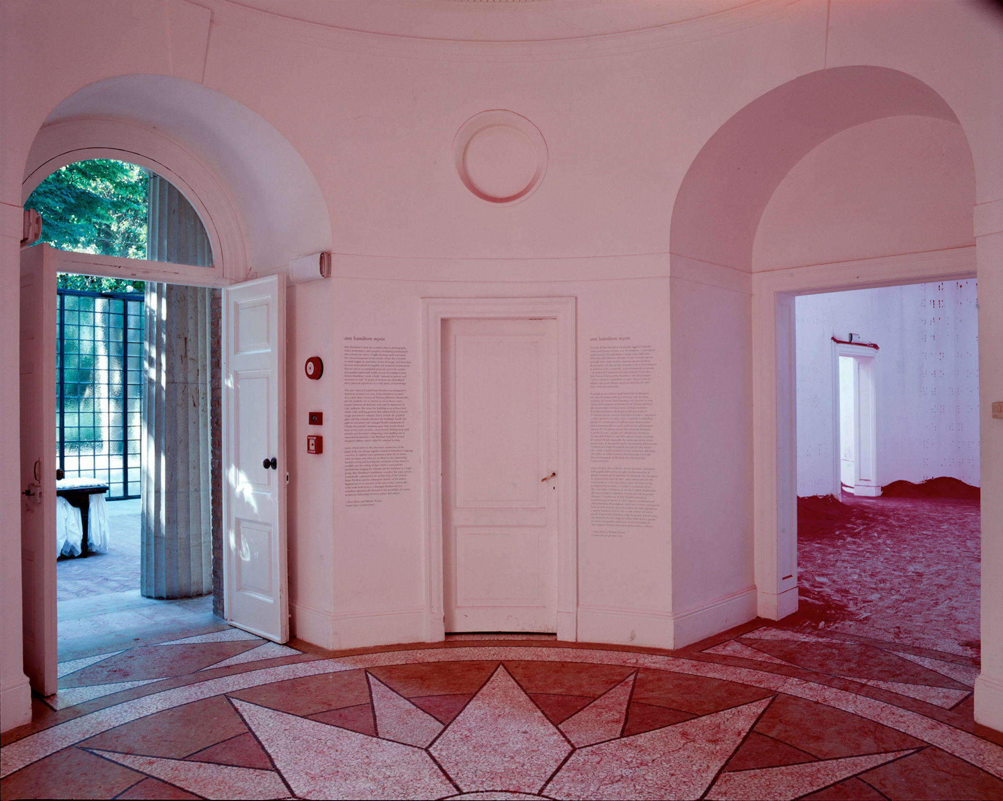 A symmetrical room with pink-tinted walls, an ornate star pattern on the floor, and two arched doorways. One doorway opens to a sunlit outdoor area, the other into another white walled room.