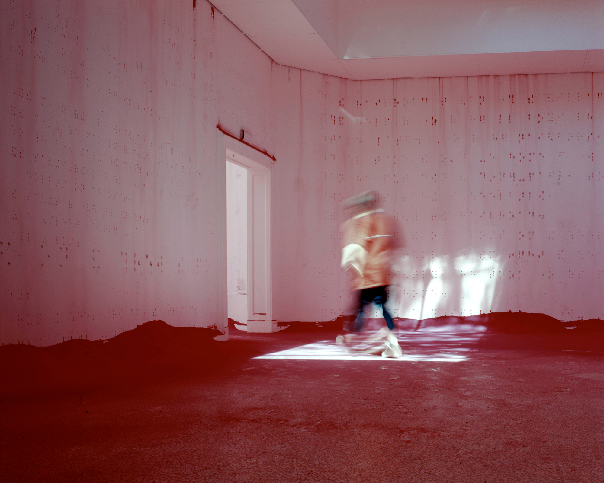 A blurred figure walks through a door in a dimly lit room with red-stained walls and red powder covered floor. Sunlight peaks in through the doorway and creates an ethereal glow on the back wall.