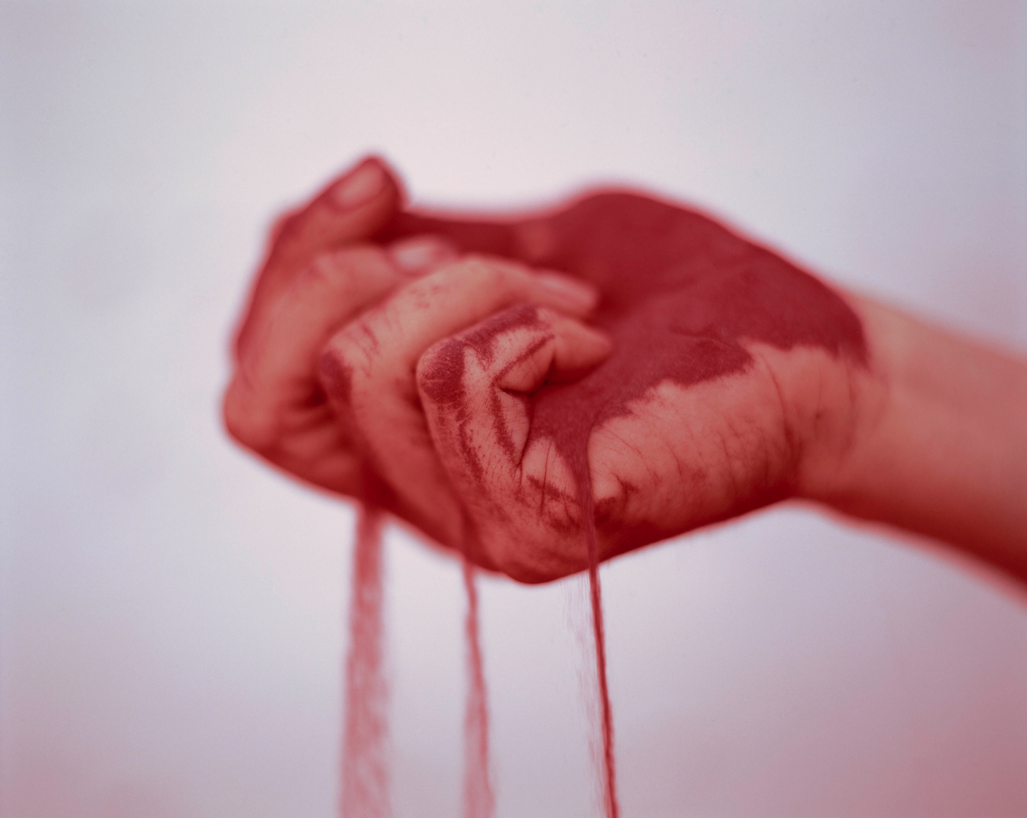 Close-up of a hand holding red sand, with grains slipping through the fingers, in front of a white wall.
