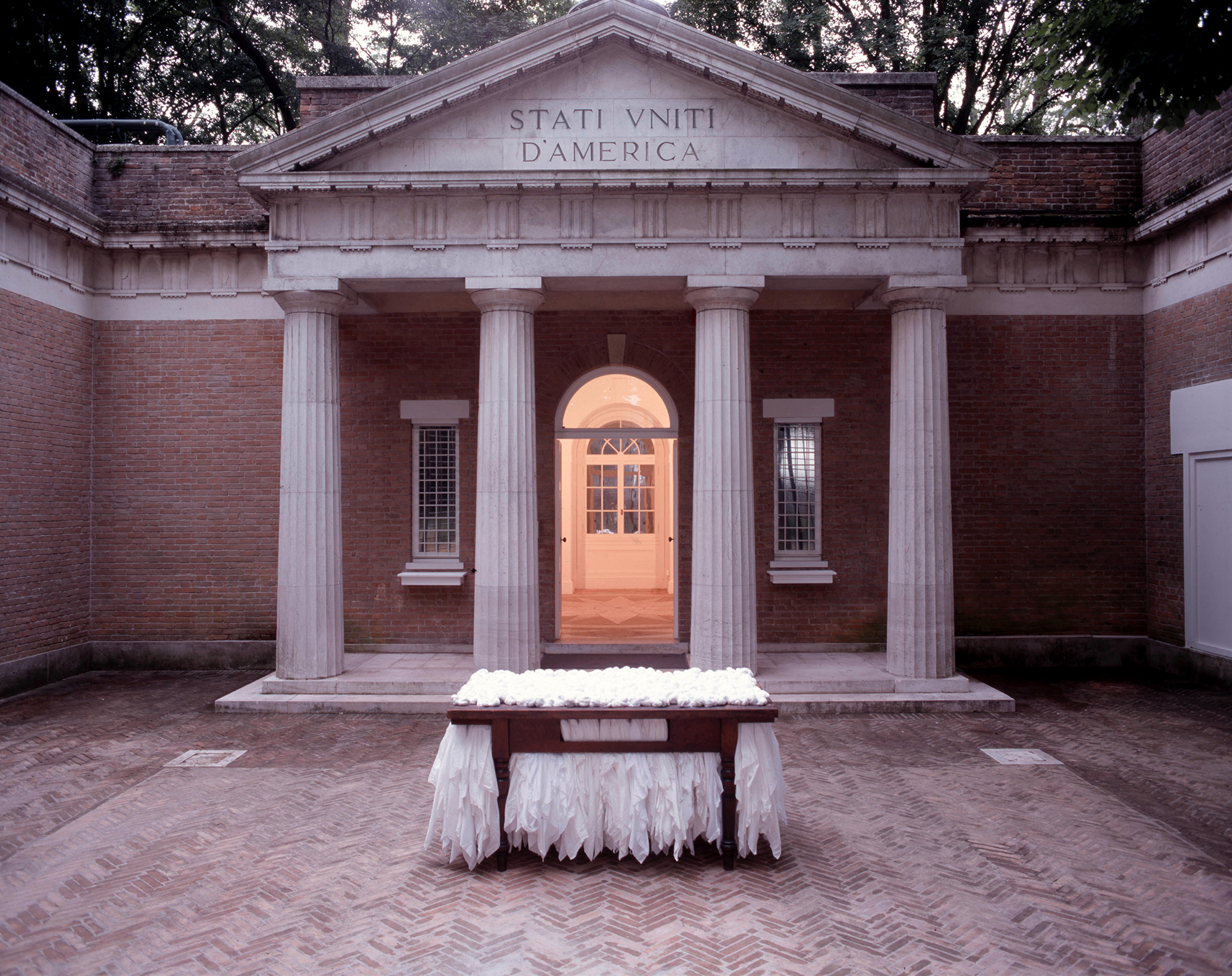 Neoclassical building with columns labeled "Stati Uniti d'America." In the foreground, a table with white fabric is set on a brick courtyard.