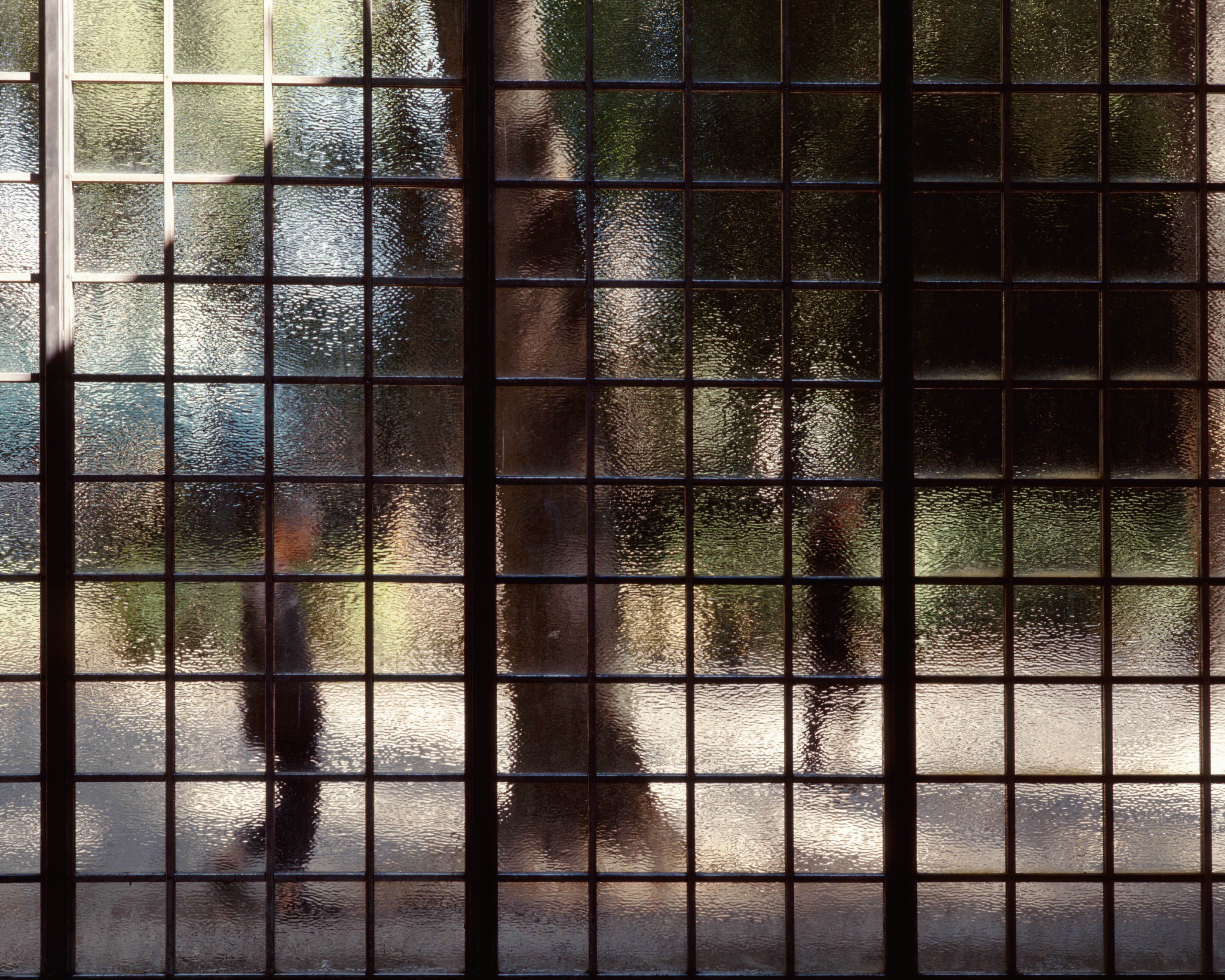 A gridded metal fence with blurry panes of glass. The blurry figure of a person walking outside in the distance can be seen through the glass.