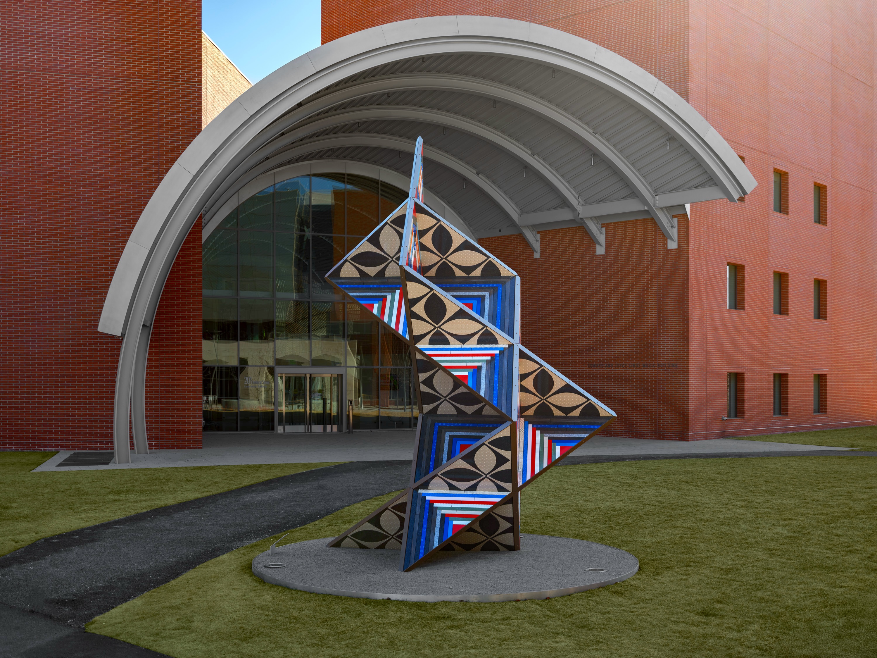 Large brightly colored geometric sculpture in the middle of a grass lawn in front of a brick building with a concrete dome awning.