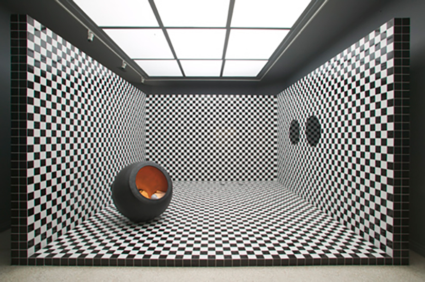 Room with black-and-white checkerboard walls and floor. A black circular object sits in the corner, creating an illusion of depth and distortion. The ceiling is a sky light with white opaque glass.