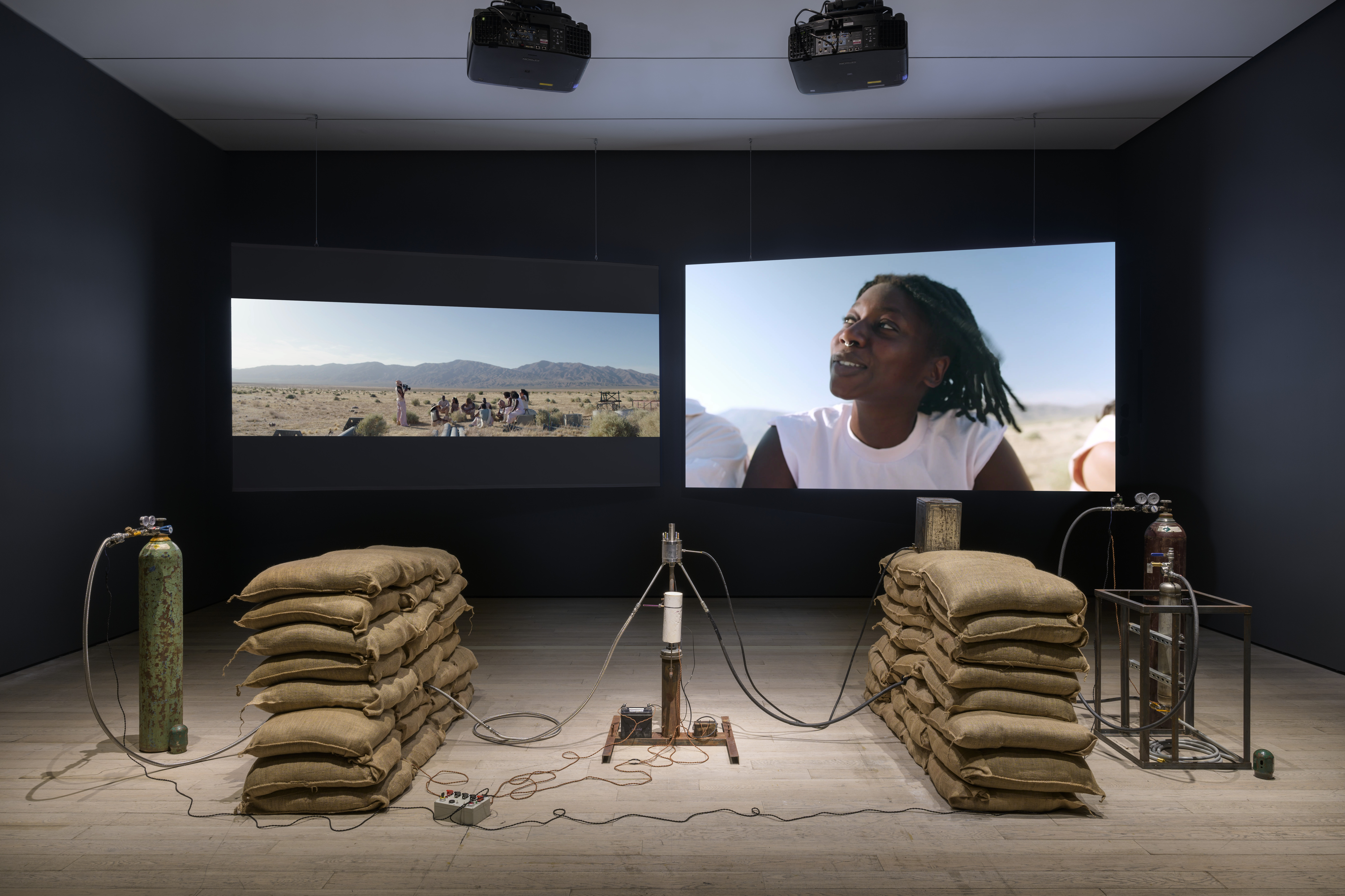 Dimly lit galleries space with two videos playing on projector screens on the back wall. Two burlap bags are in the middle of the exhibition space.