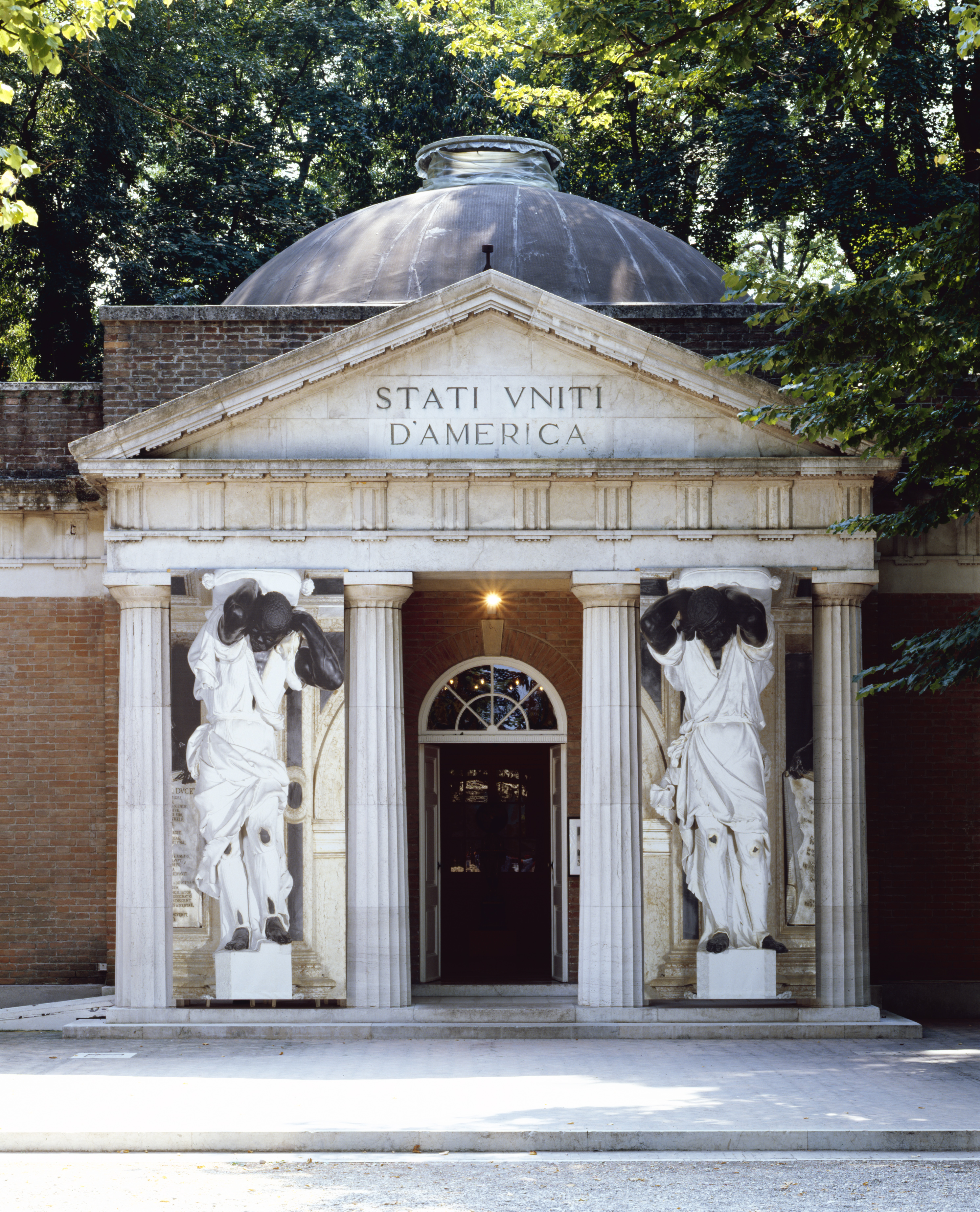 A classical stone building featuring tall columns supporting a grand entrance. There are two human figure statues on either side of the doorway. The text on the building facade reads, "STATI VNITI D'AMERICA"