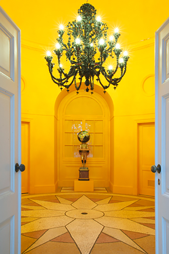 A grand black chandelier illuminates a vibrant orange room with paneled doors. A geometric mosaic floor pattern centers the space, creating an elegant ambiance.
