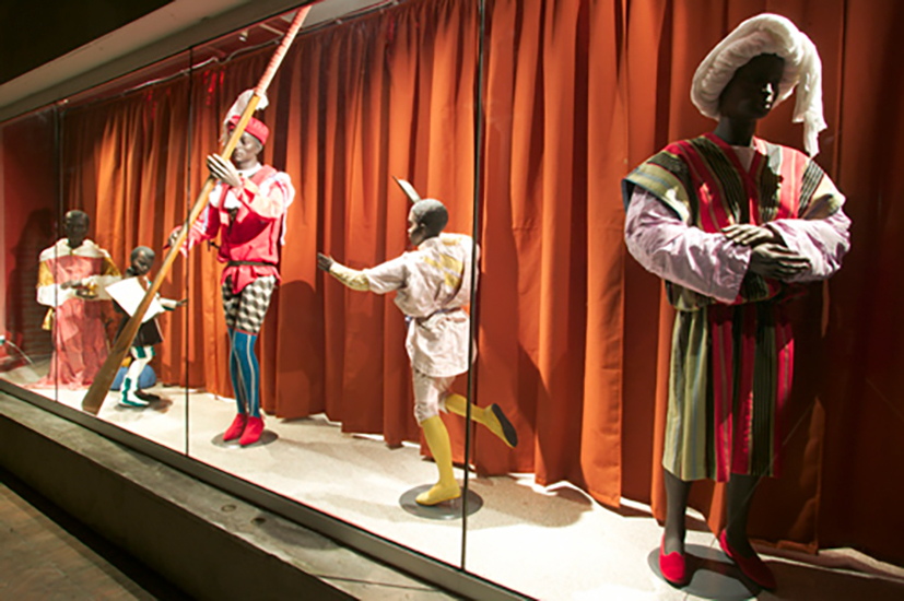 Mannequins dressed in colorful, historical costumes, each striking dynamic poses, are displayed against a redish orange curtain backdrop in a glass case.