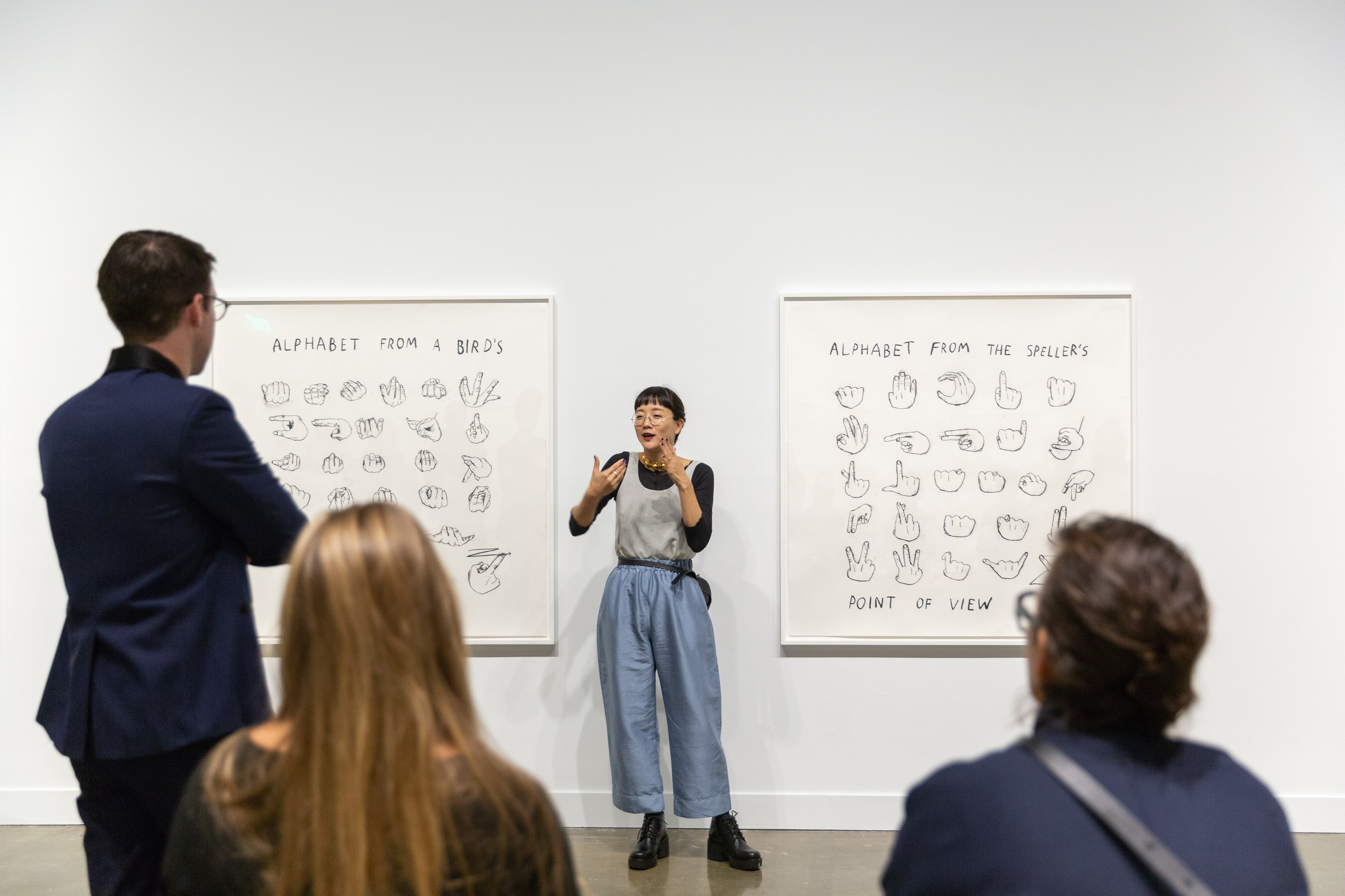 Three visitors attending a gallery talk with their backs to the camera are looking at the central speaker who is signing in ASL.
