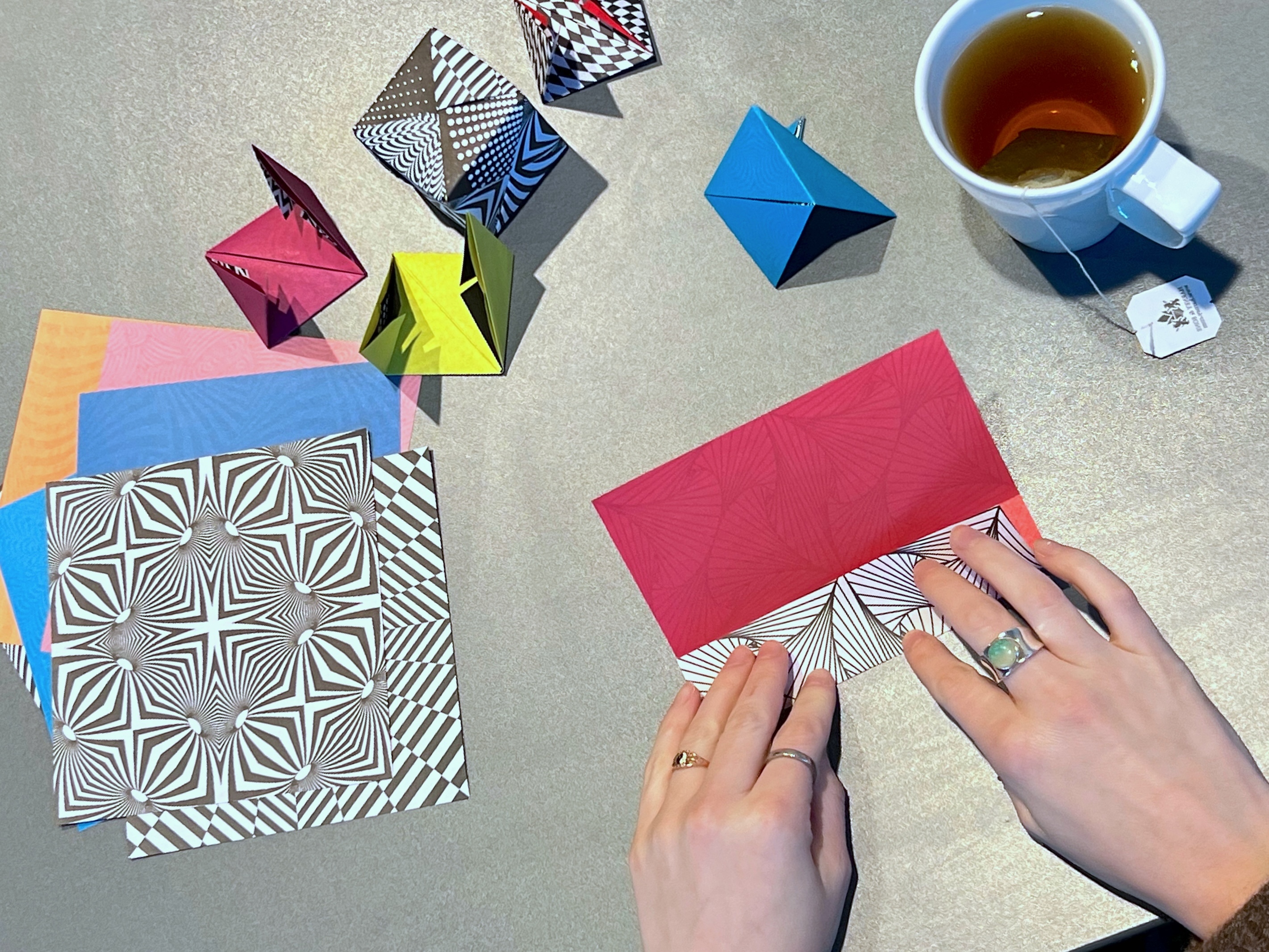 Overhead view of two hands folding colorful geometric paper. There is a white mug of tea to the left side and a stack of unfolded paper to the right.