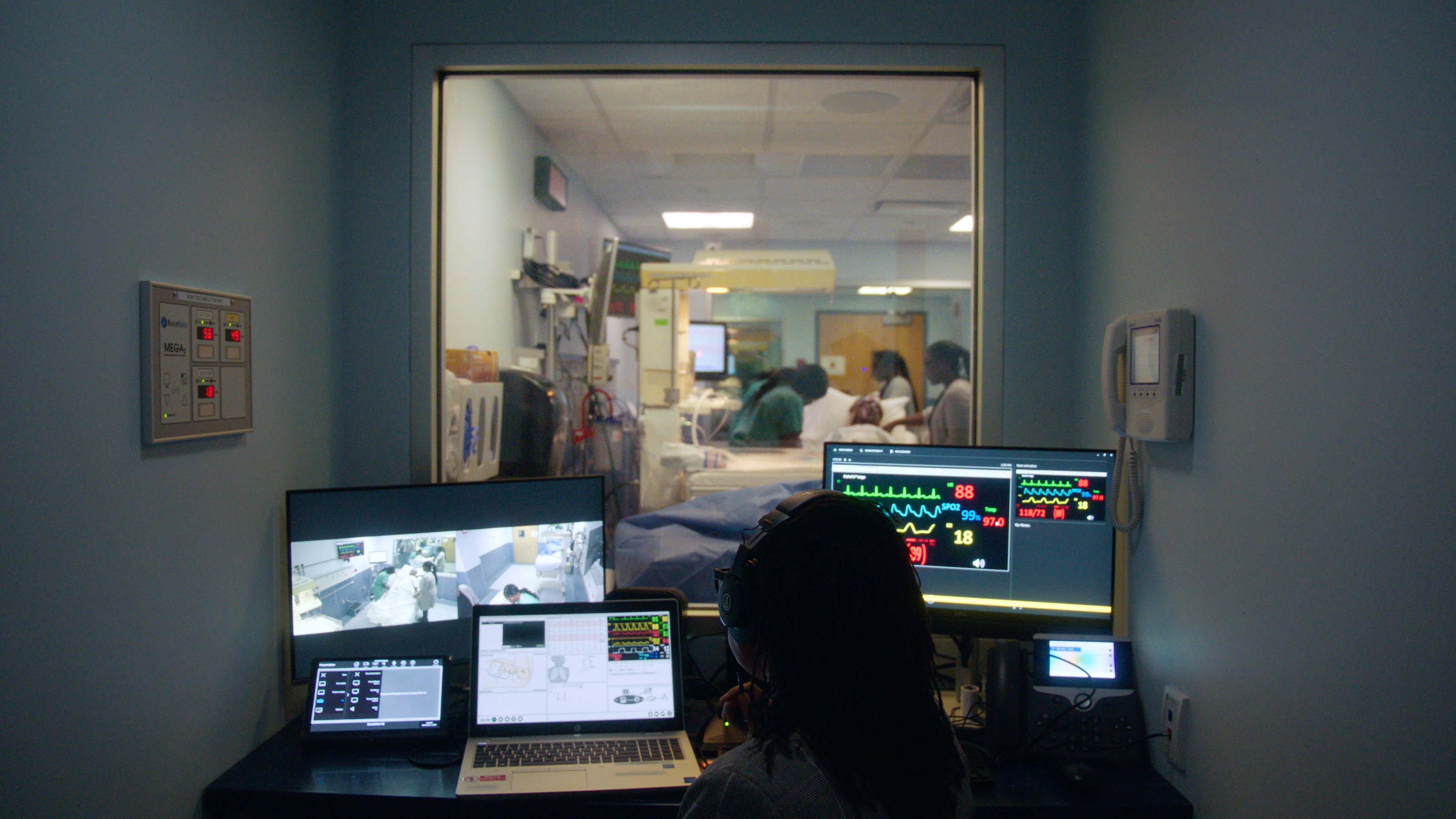 In a small dark control room, a person sits in front of multiple computer monitors while observing people in an adjoining patient room via an interior window.
