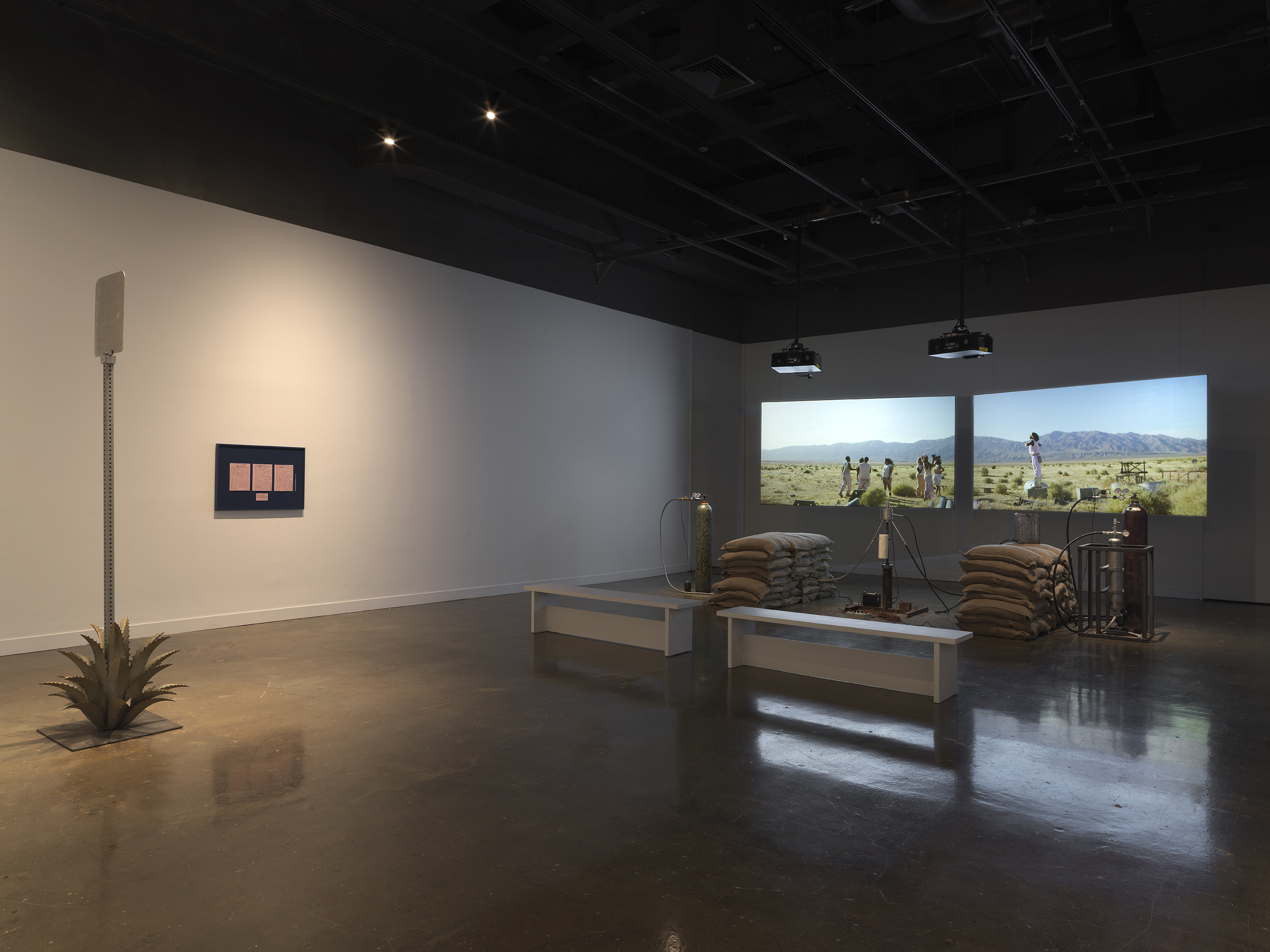 Inside a dimly lit gallery space, two projections sit behind a rocket test installation containing sandbags and canisters. In the foreground to the left is a metal sign inside an agave plant and a blue frame with pink paper mounted.