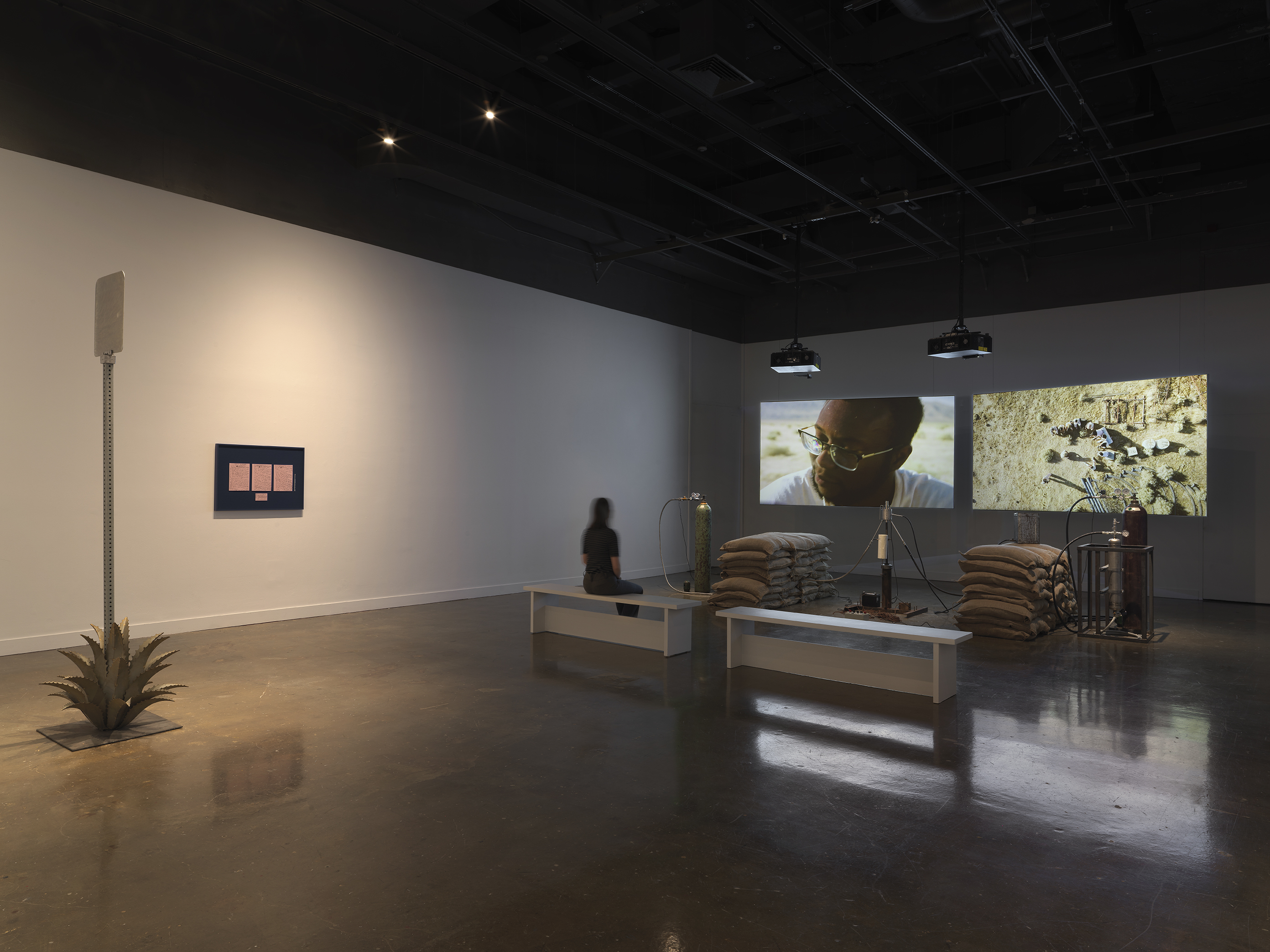 Inside a dimly lit gallery space, a long figure sits on a bench watching two projections sitting behind a rocket test installation containing sandbags and canisters. In the foreground to the left is a metal sign inside an agave plant and a blue frame with pink paper mounted.