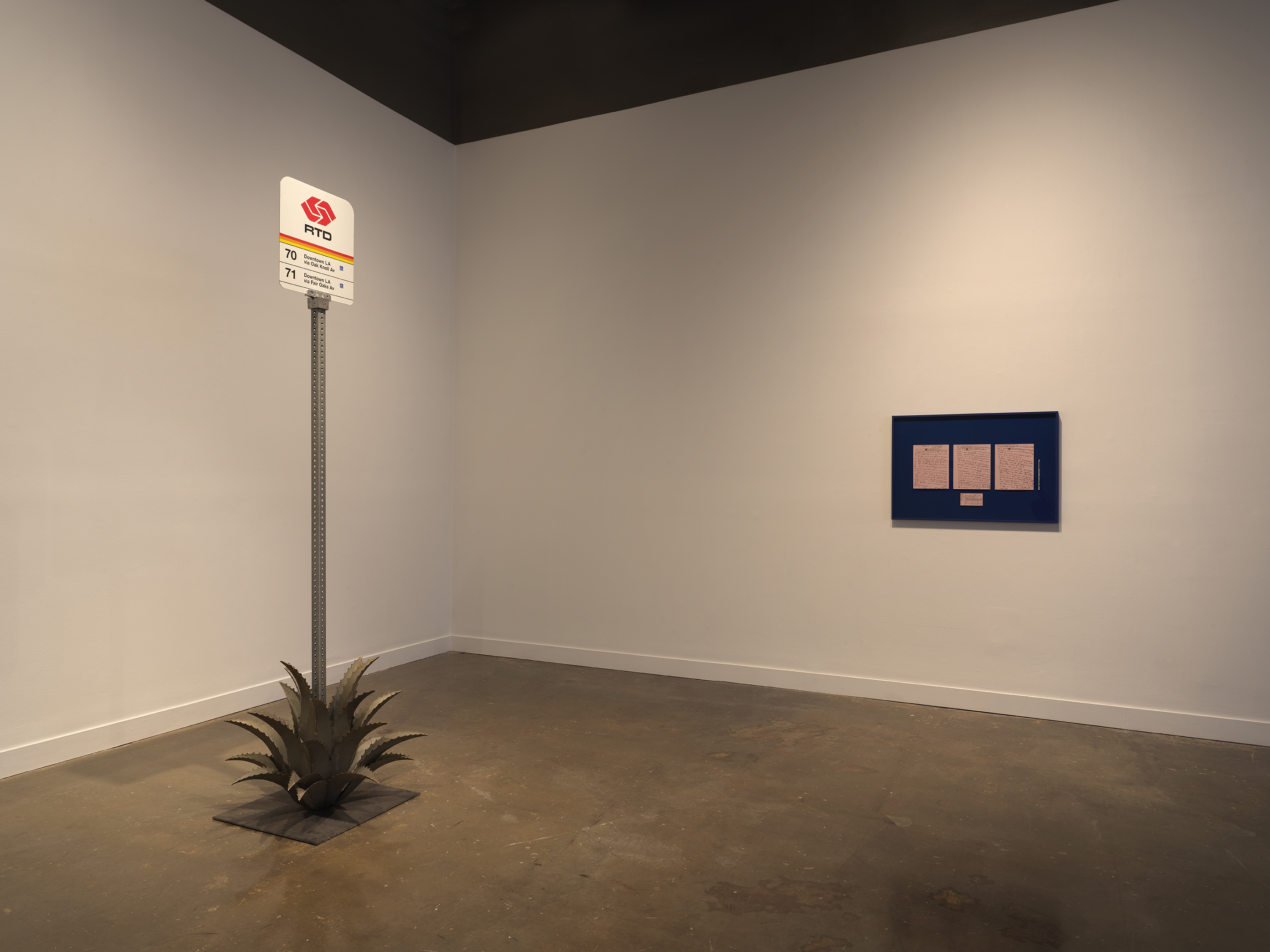 In a white gallery space, a Pasadena bus stop sign sits amidst a metal agave plant and on the right a blue frame is hung on the wall with 4 pink papers mounted inside.