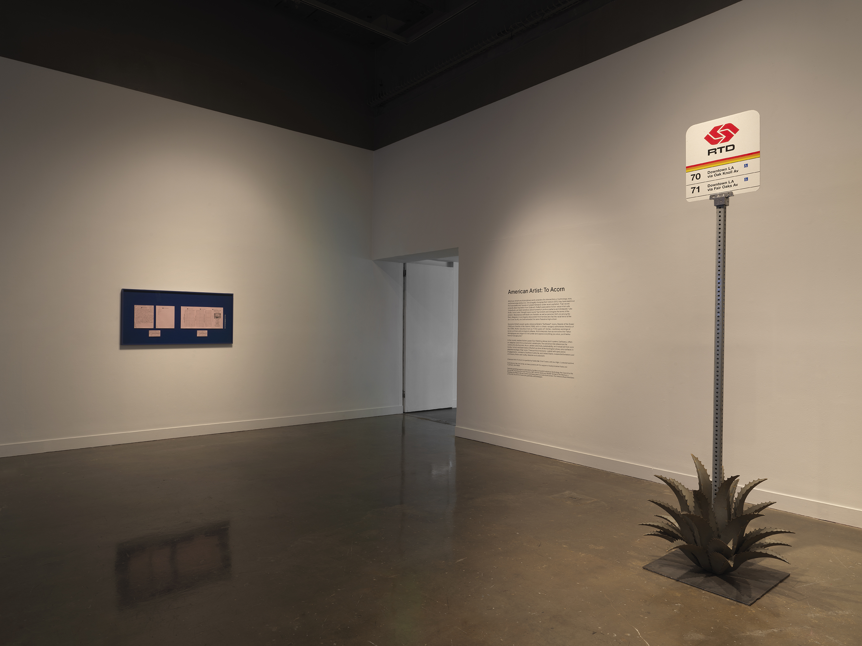 In a white gallery space, a Pasadena bus stop sign sits amidst a metal agave plant and on the left, a blue frame is hung on the wall with 5 pink papers mounted inside.