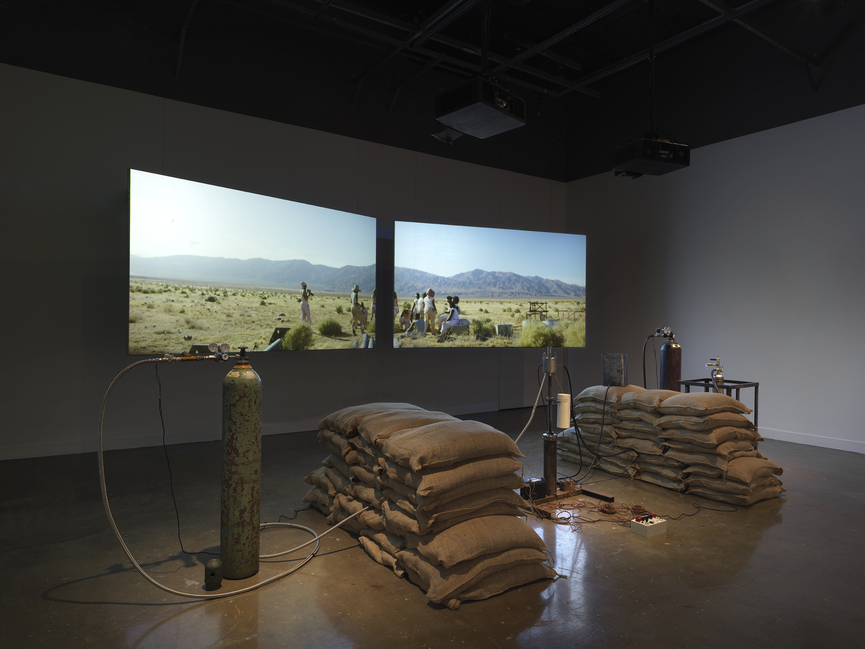 An installation of a rocket test containing sandbags, wiring, and canisters rest in a gallery space in front of two projections depicting people sitting in a desert landscape.