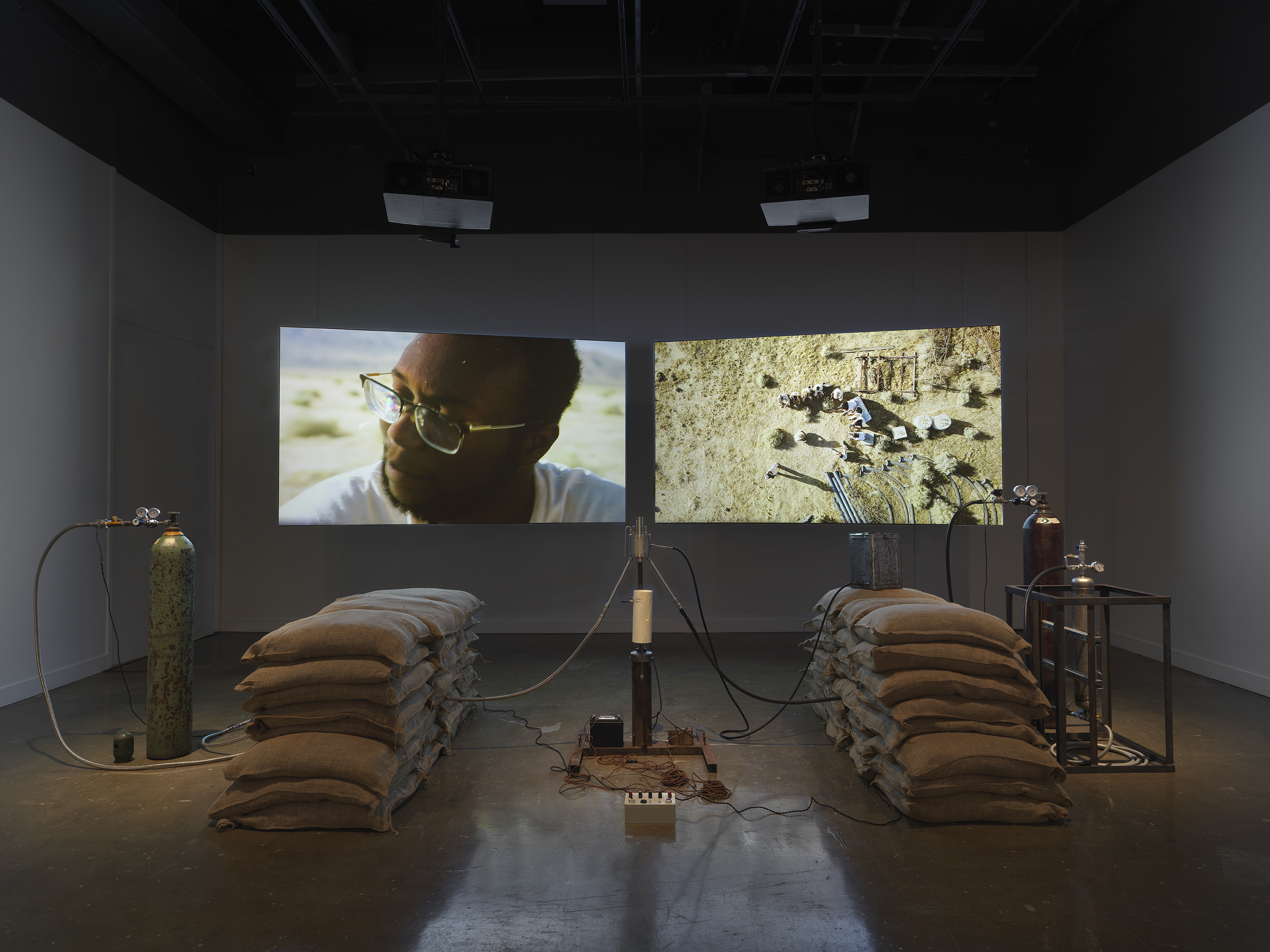 An installation of a rocket test containing sandbags, wiring, and canisters rest in a gallery space in front of two projections depicting people sitting in a desert landscape and a closeup of a man with dark skin tone gazing outwards.