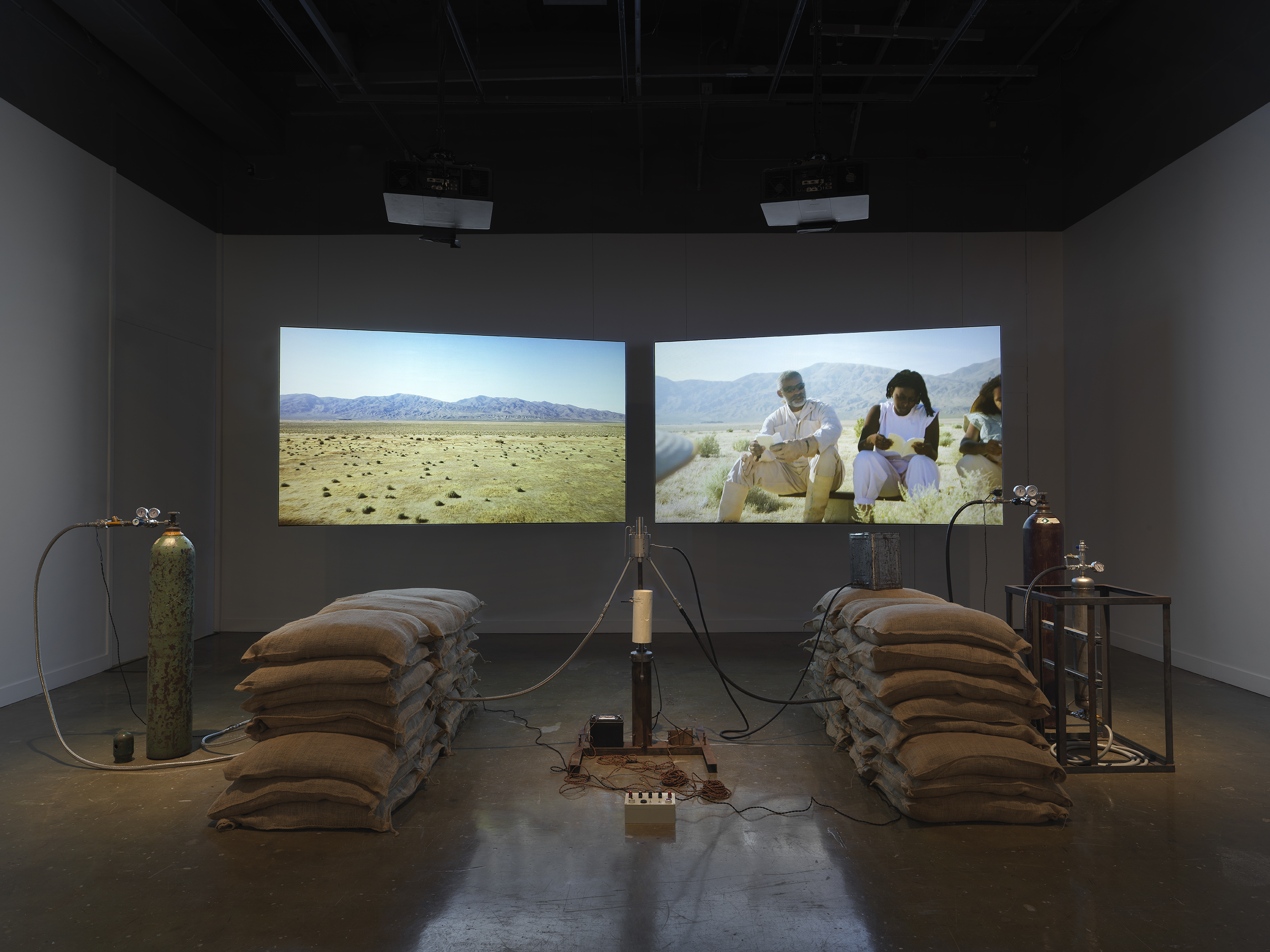 An installation of a rocket test containing sandbags, wiring, and canisters rest in a gallery space in front of two projections depicting people sitting in a desert landscape.
