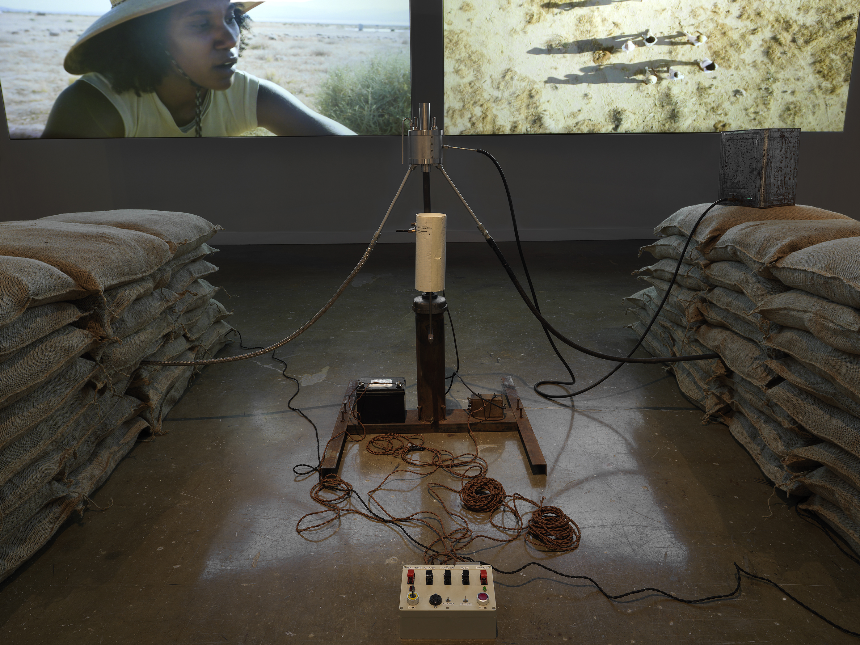 Part of an installation of a rocket test containing sandbags and wiring rest in a gallery space in front of two projections depicting people in a desert landscape.