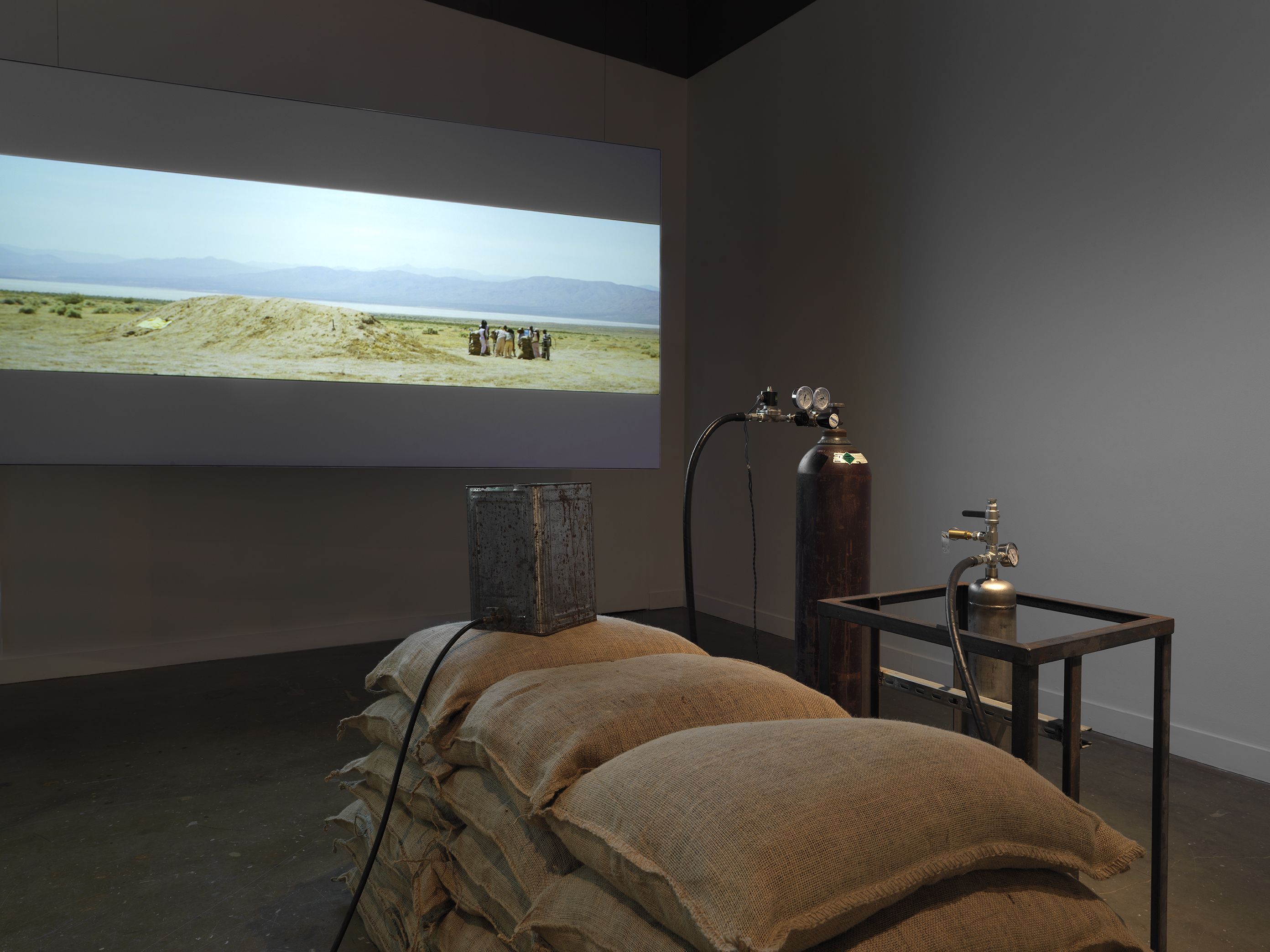 Part of an installation of a rocket test containing sandbags and canisters resting in a gallery space in front of a projection depicting people standing in a desert landscape.