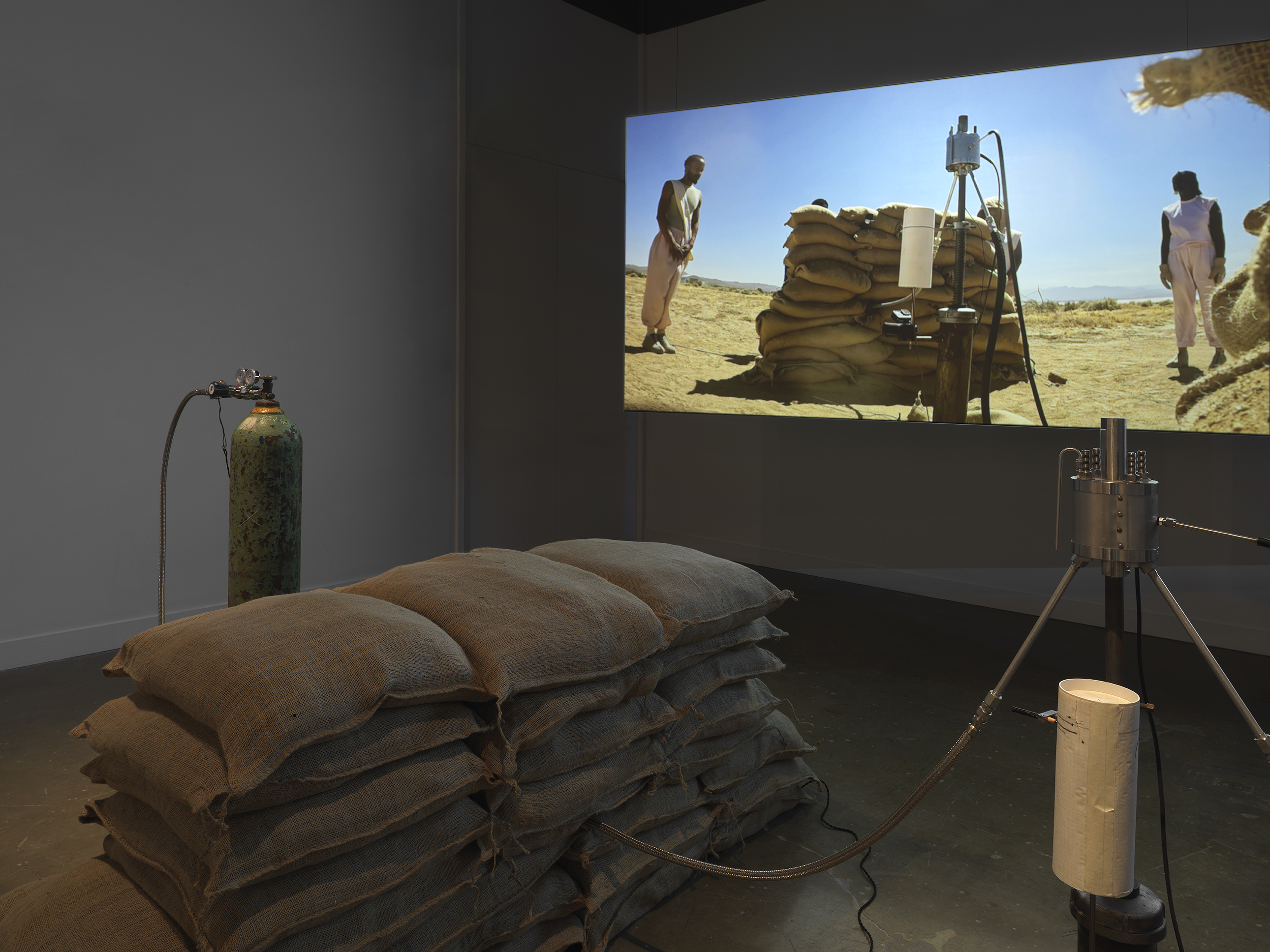 Part of an installation of a rocket test containing sandbags and canisters rest in a gallery space in front of a projection depicting people standing in a desert landscape looking at a similar rocket test setup.