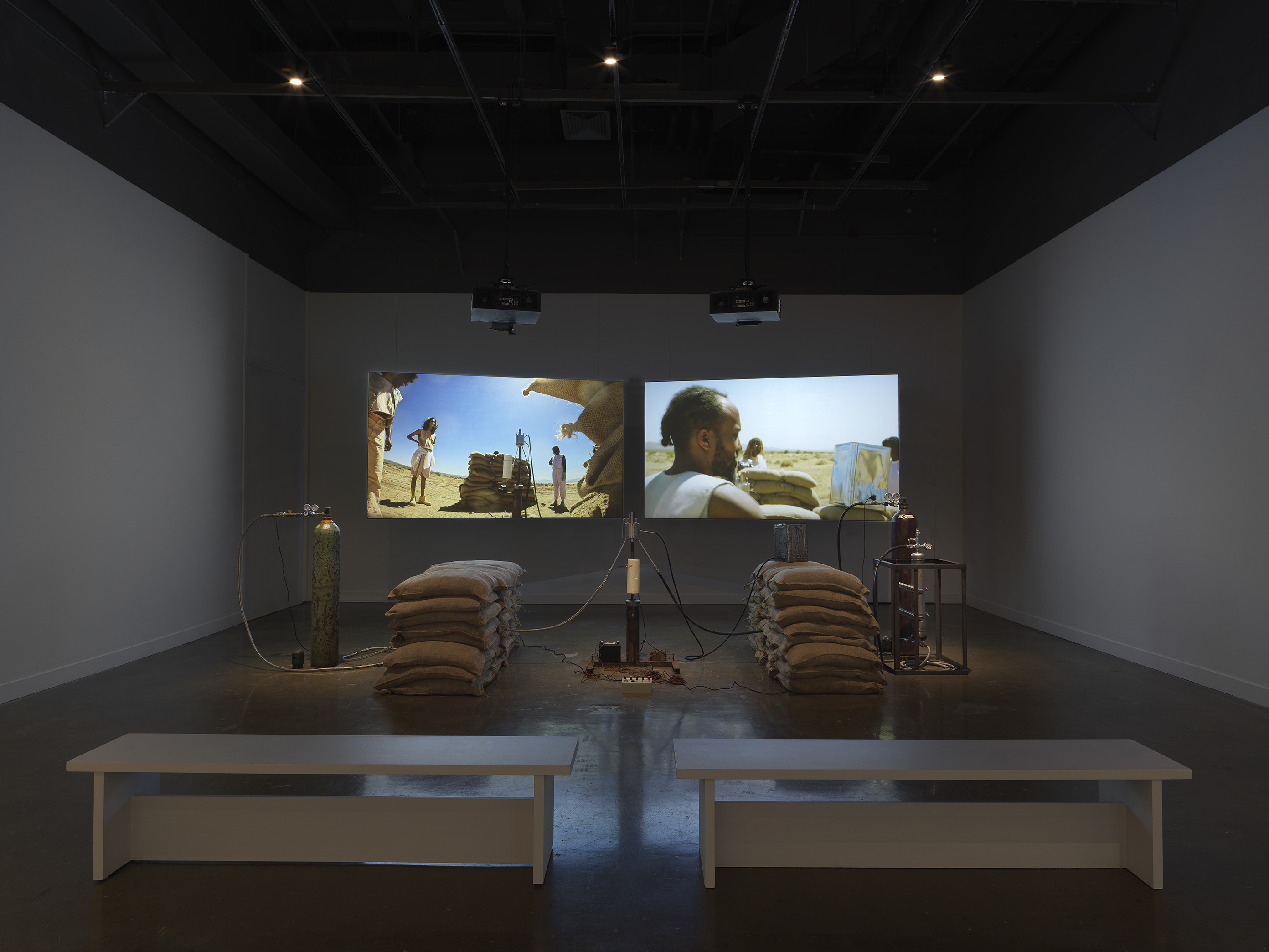 An installation of a rocket test containing sandbags, wiring, and canisters rest in a gallery space in front of two projections depicting people standing in a desert landscape around a similar rocket test set-up. Two white benches sit in the foreground.