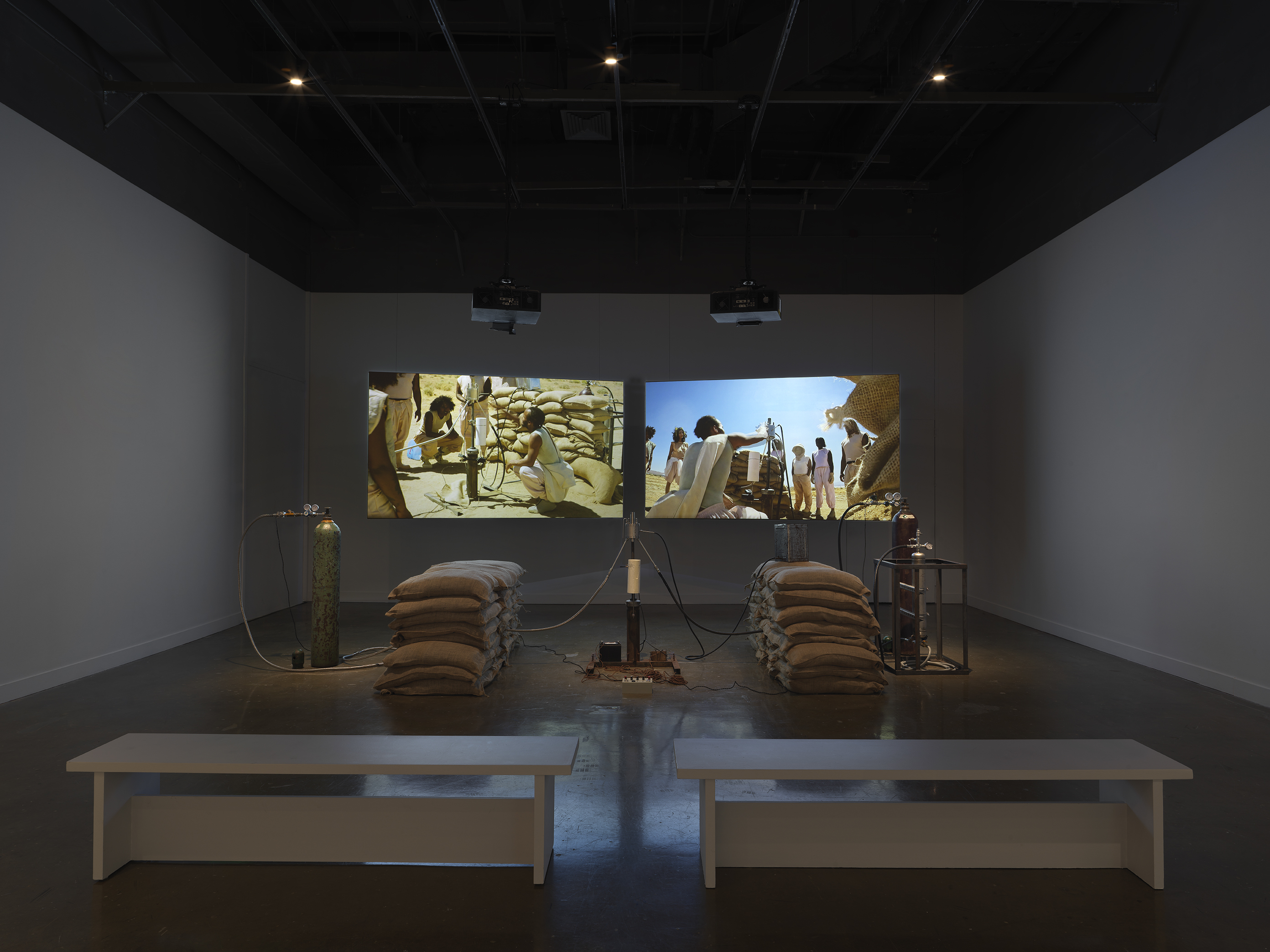 An installation of a rocket test containing sandbags, wiring, and canisters resting in a gallery space in front of two projections depicting people standing in a desert landscape around a similar rocket test set-up. Two white benches sit in the foreground.