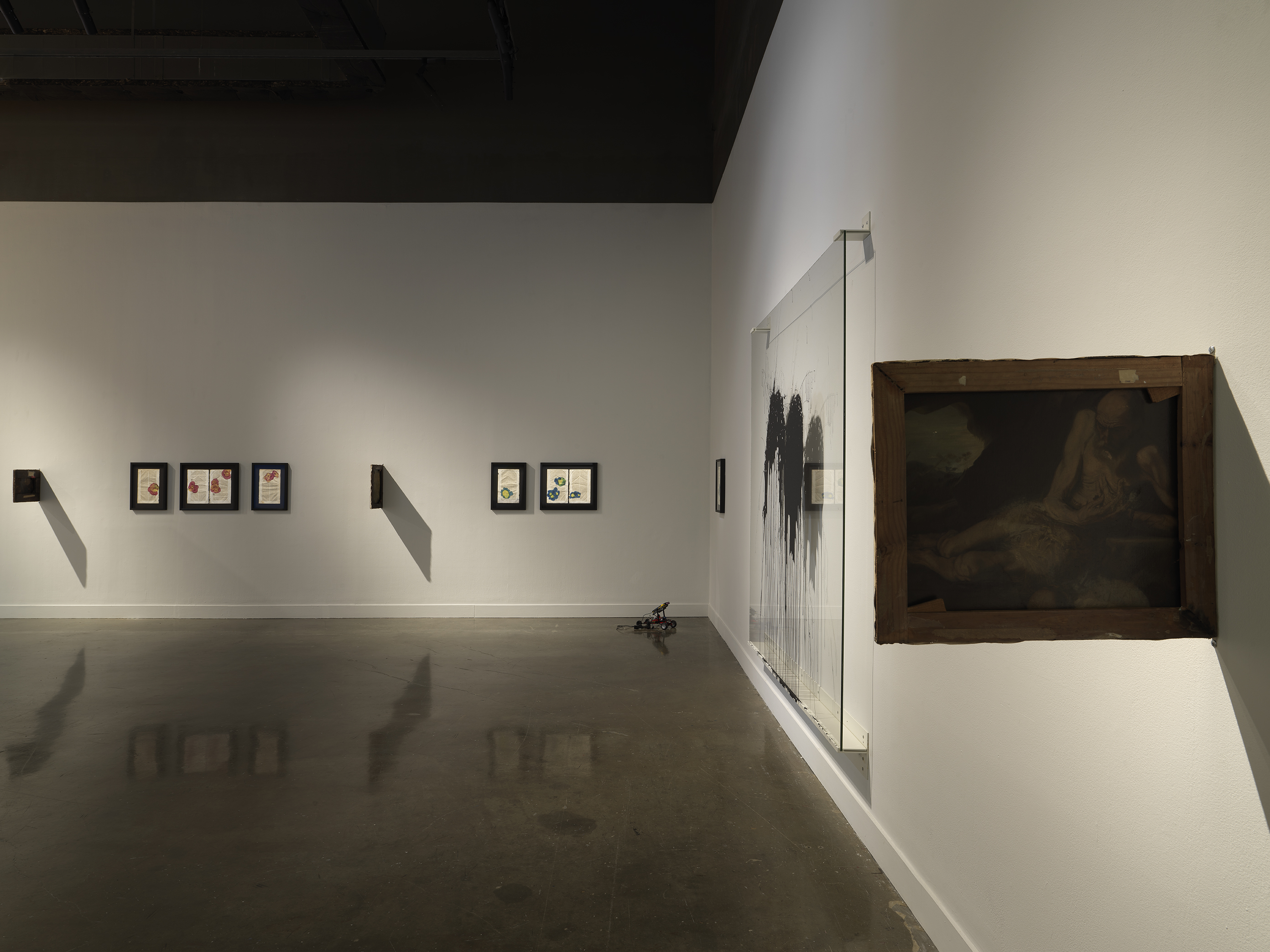 A series of black framed pages of text with colorful botanical ink blots hang in a row on white walls in a dimly lit gallery. A small lego robot sits on the floor in the corner of the space. On the right side of the frame, the artwork closest to the viewer hangs perpendicular to the wall.