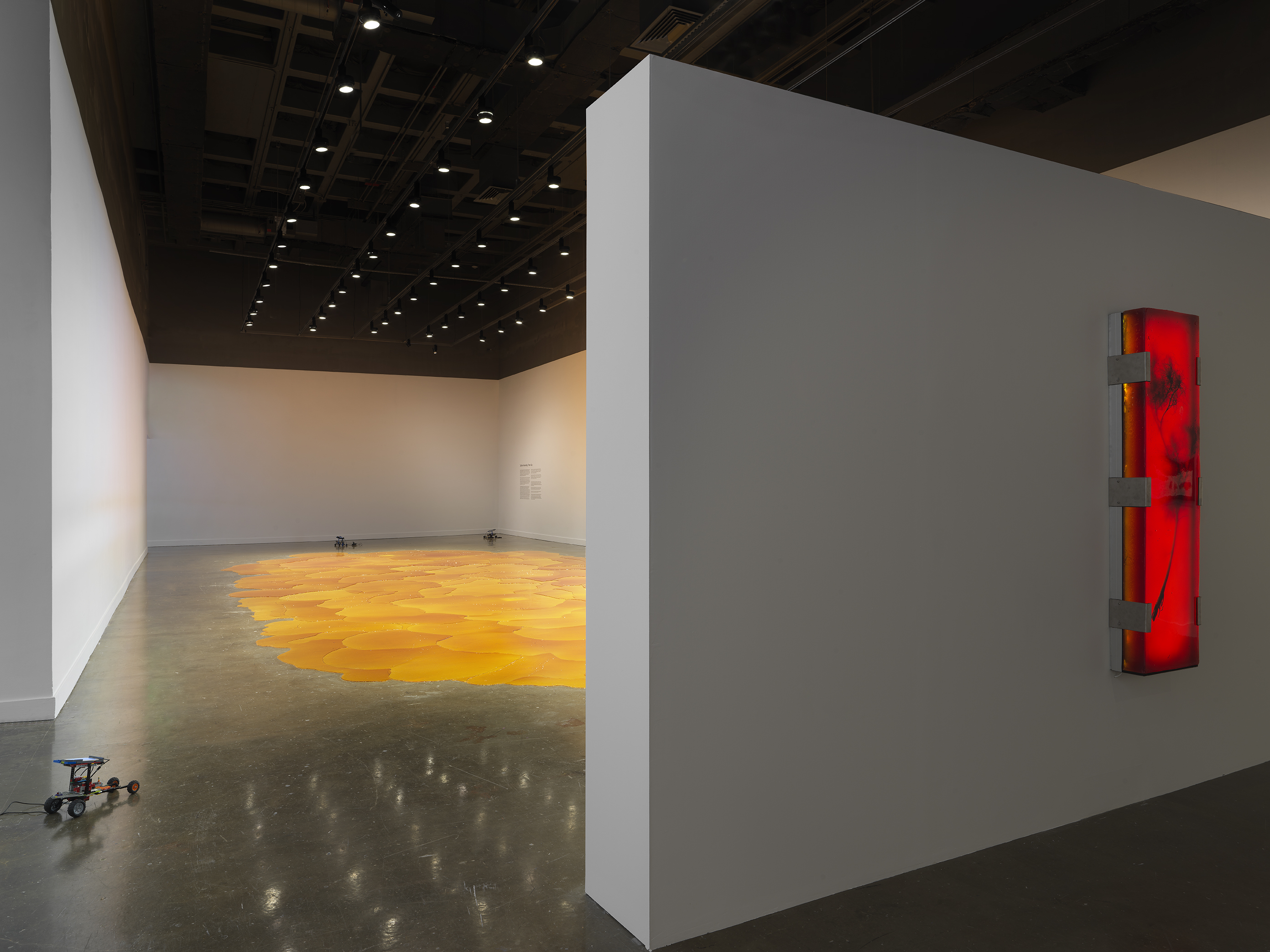 Gallery with an expansive, vibrant orange pool of melted pine rosin that spreads across the floor. Small lego robots are placed throughout the space. A large rectangular illuminated red-orange artwork hangs on a wall on the right side of the frame.