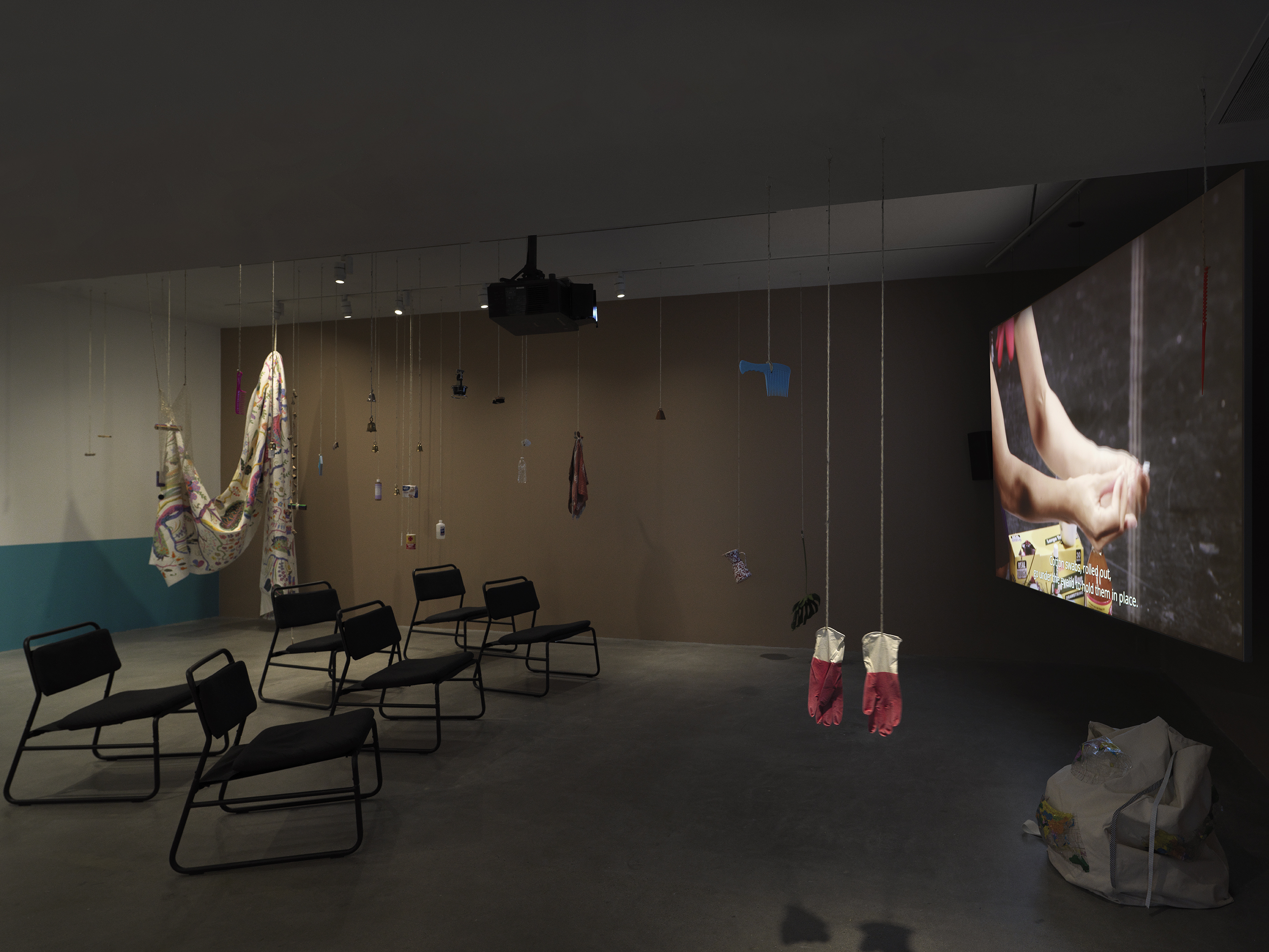 Dark open gallery with a video projected on a large standing screen on the right side of the frame and six black chairs in two rows in the center of the room. A tan canvas bag sits on the floor to the right of the screen. There is a collection of everyday items hanging from strings throughout the space. Two bright red rubber gloves hang in the foreground.