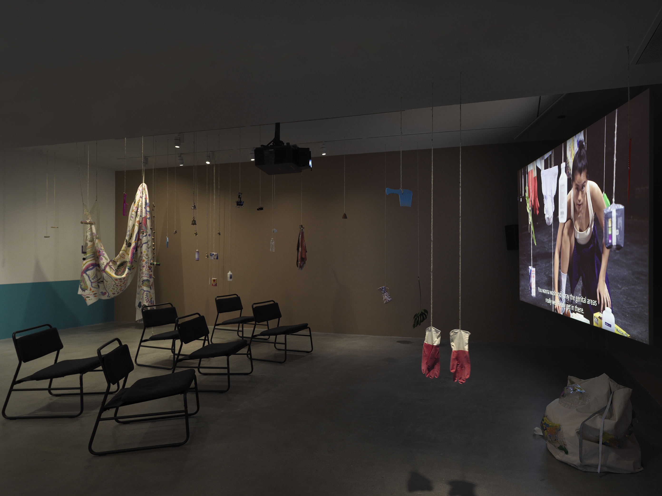 Dark open gallery with a video projected on a large standing screen on the right side of the frame and six black chairs in two rows in the center of the room. A tan canvas bag sits on the floor to the right of the screen. There is a collection of everyday items hanging from strings throughout the space. Two bright red rubber gloves hang in the foreground.