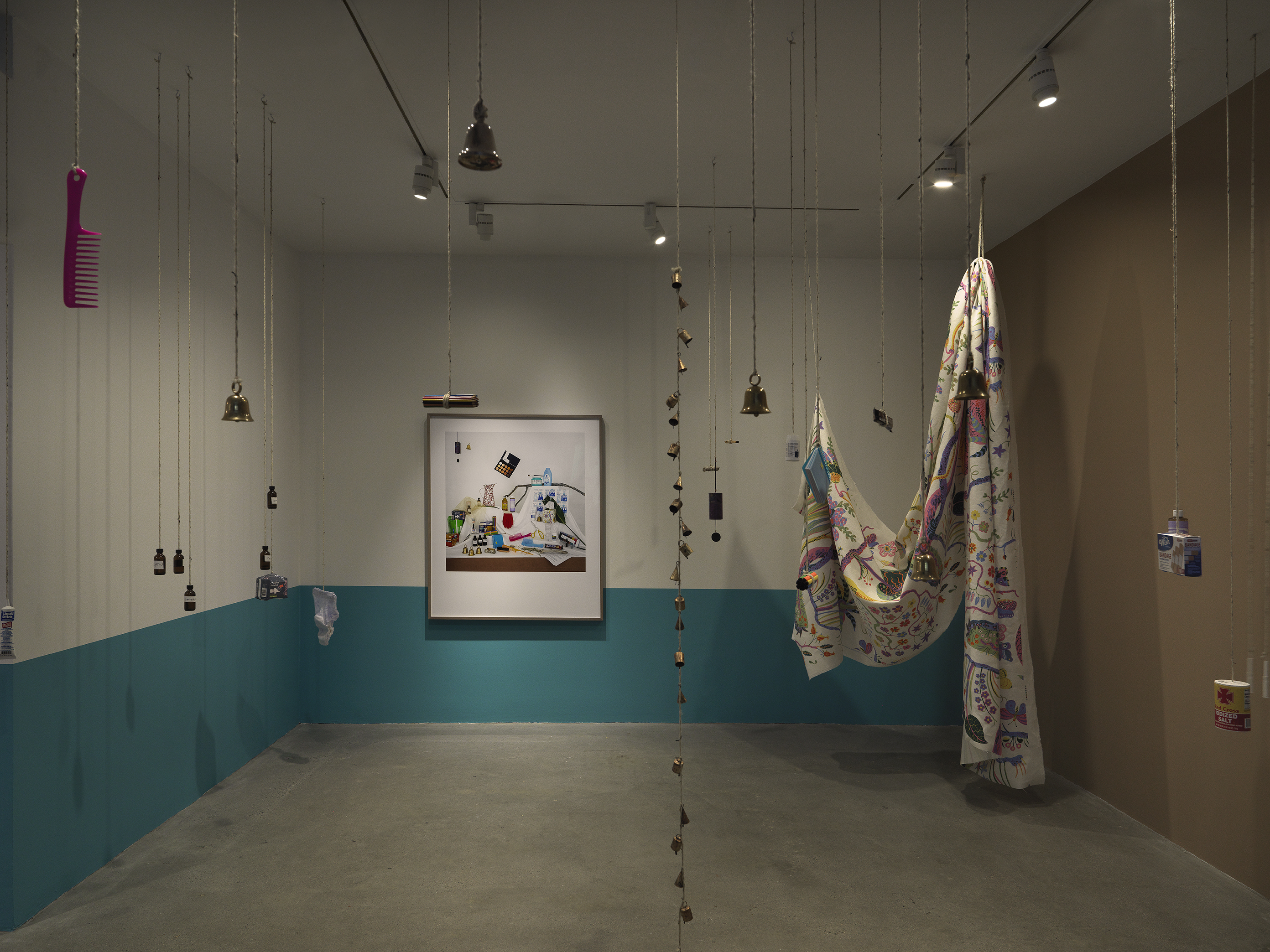 A dimly lit white walled gallery with the bottom half of the wall painted turquoise. A collection of everyday items hanging from strings throughout the space and a large black framed color photograph hangs on the back wall. A large printed sheet of fabric hangs on the right side of the frame.