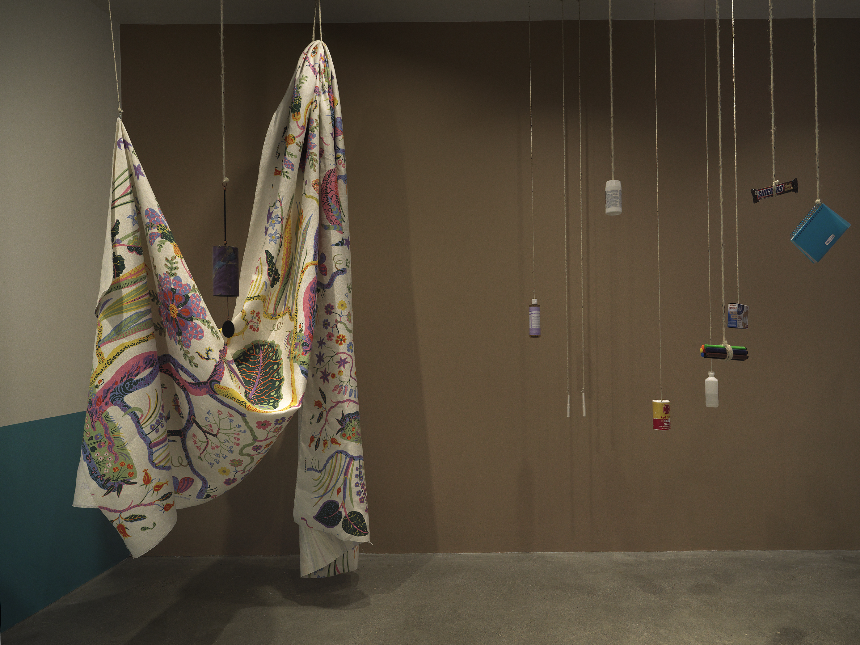 A dimly lit gallery with brown walls and a collection of everyday items hanging from strings throughout the space. A large printed sheet of fabric hangs on the left side of the frame.
