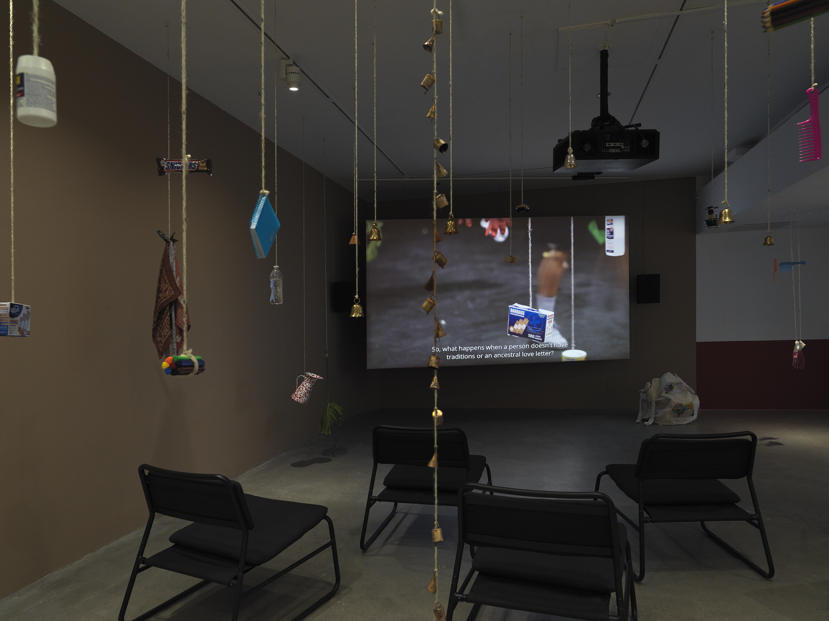 Dark open gallery with a video projected on a large standing screen in the back of the space. There is a collection of everyday items hanging from strings throughout the gallery. A tan canvas bag sits on the floor to the right of the screen and there is a row of chairs in the foreground.