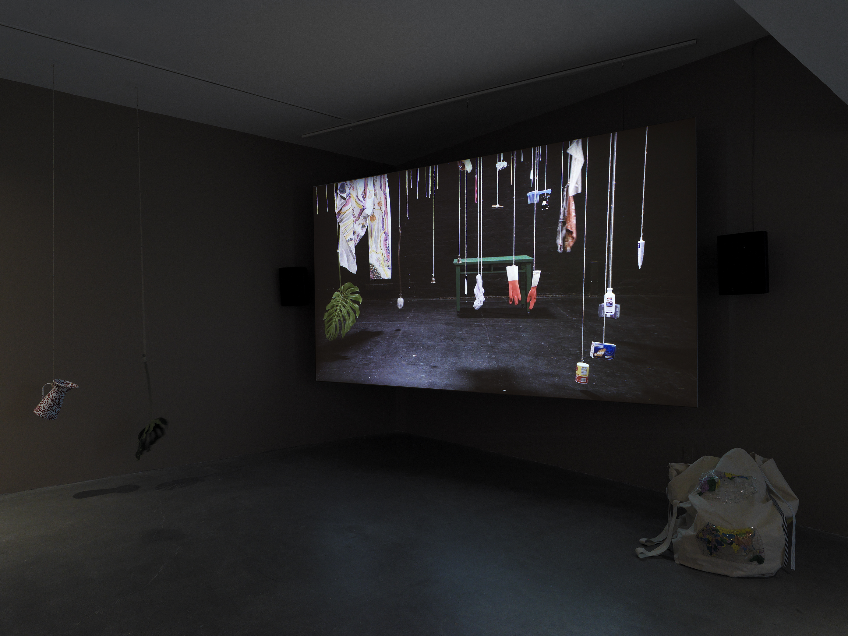 Dark open gallery with a video projected on a large standing screen in the back of the space. There is a collection of everyday items hanging from strings throughout the gallery. A tan canvas bag sits on the floor to the right of the screen.