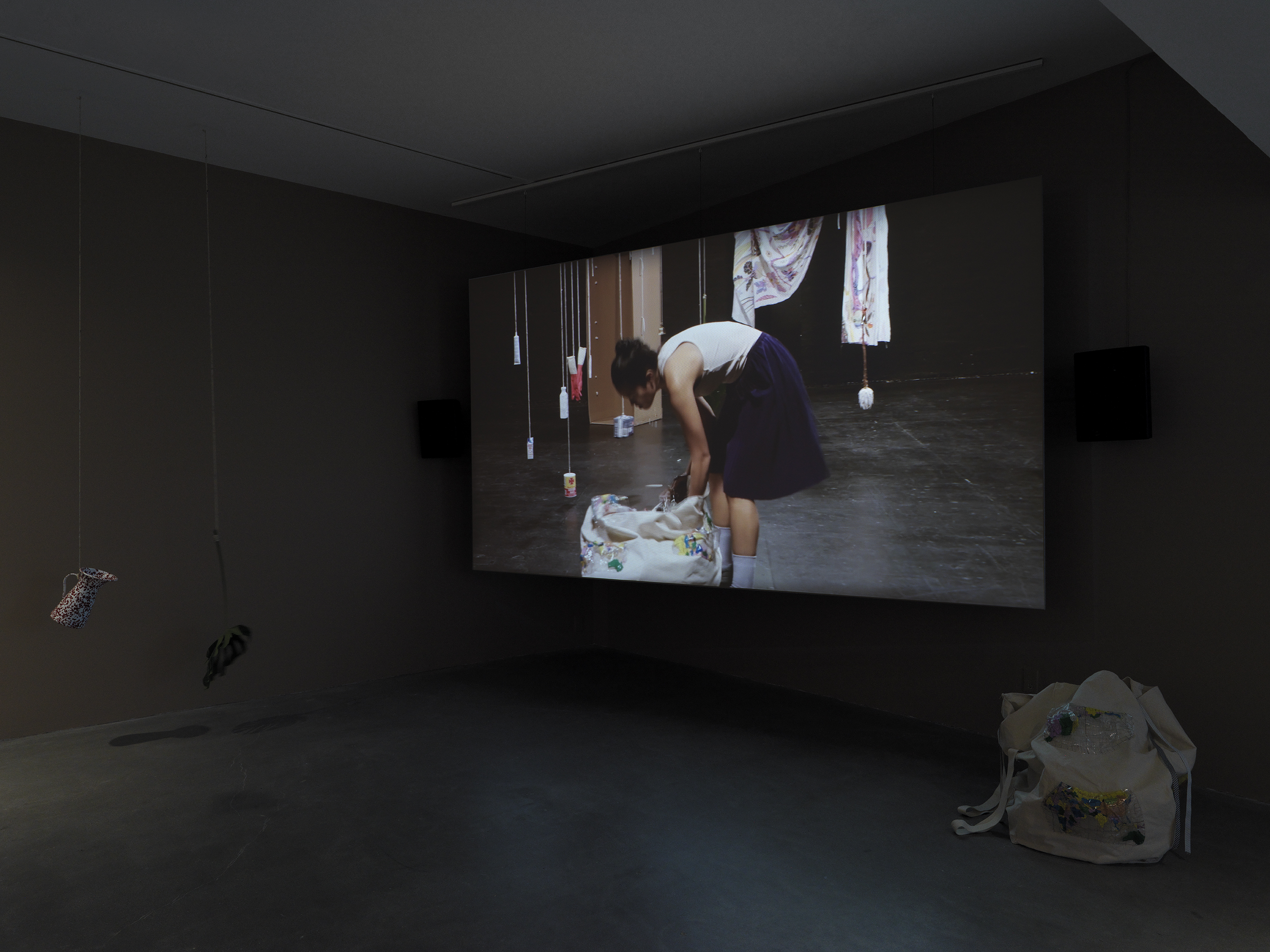 Dark open gallery with a video projected on a large standing screen in the back of the space. There is a collection of everyday items hanging from strings throughout the gallery. A tan canvas bag sits on the floor to the right of the screen.