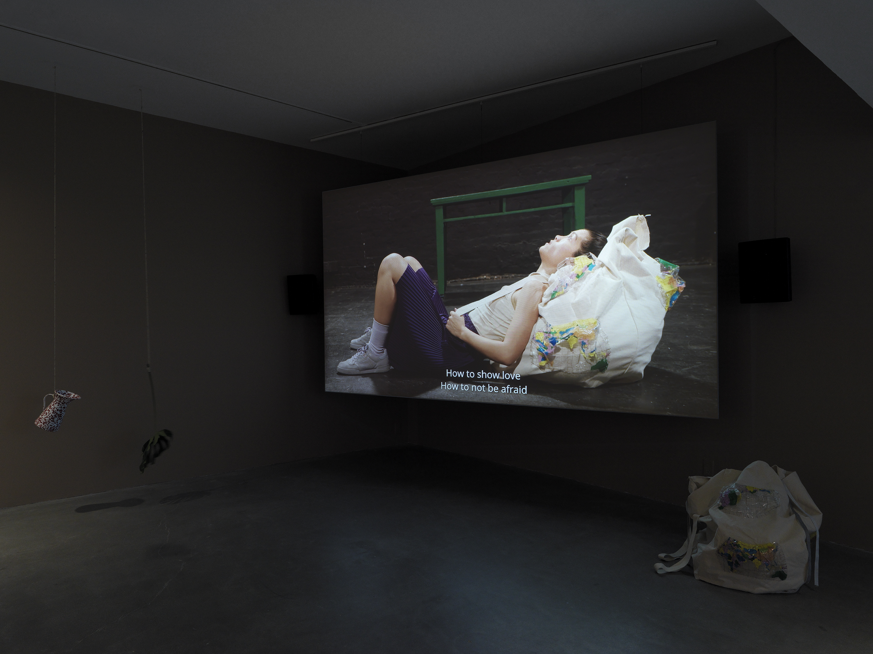Dark open gallery with a video projected on a large standing screen in the back of the space. There is a collection of everyday items hanging from strings throughout the gallery. A tan canvas bag sits on the floor to the right of the screen.