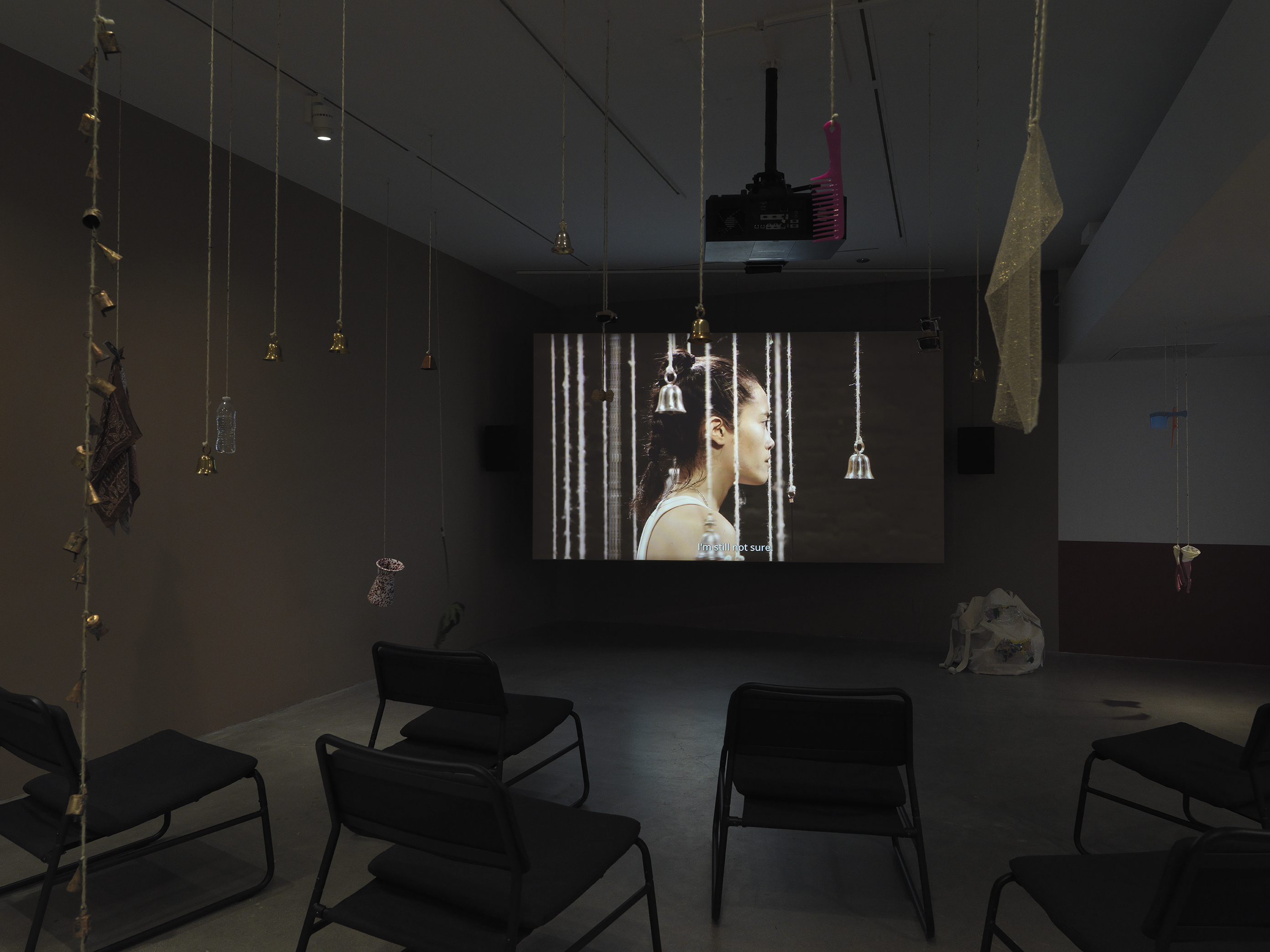 Dark open gallery with a video projected on a large standing screen in the back of the space. There is a collection of everyday items hanging from strings throughout the gallery. A tan canvas bag sits on the floor to the right of the screen and there is a row of chairs in the foreground.