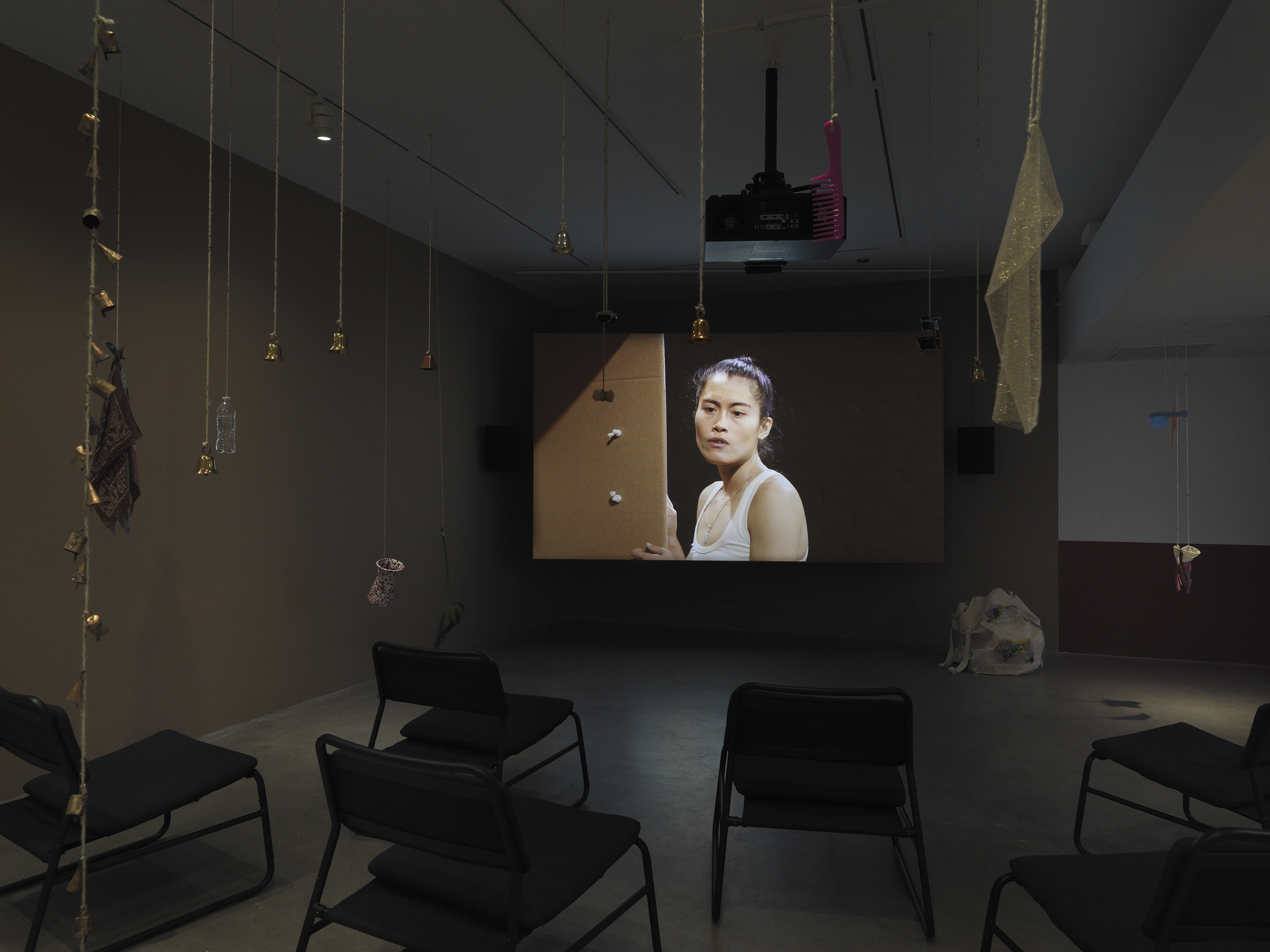 Dark open gallery with a video projected on a large standing screen in the back of the space. There is a collection of everyday items hanging from strings throughout the gallery. A tan canvas bag sits on the floor to the right of the screen and there is a row of chairs in the foreground.