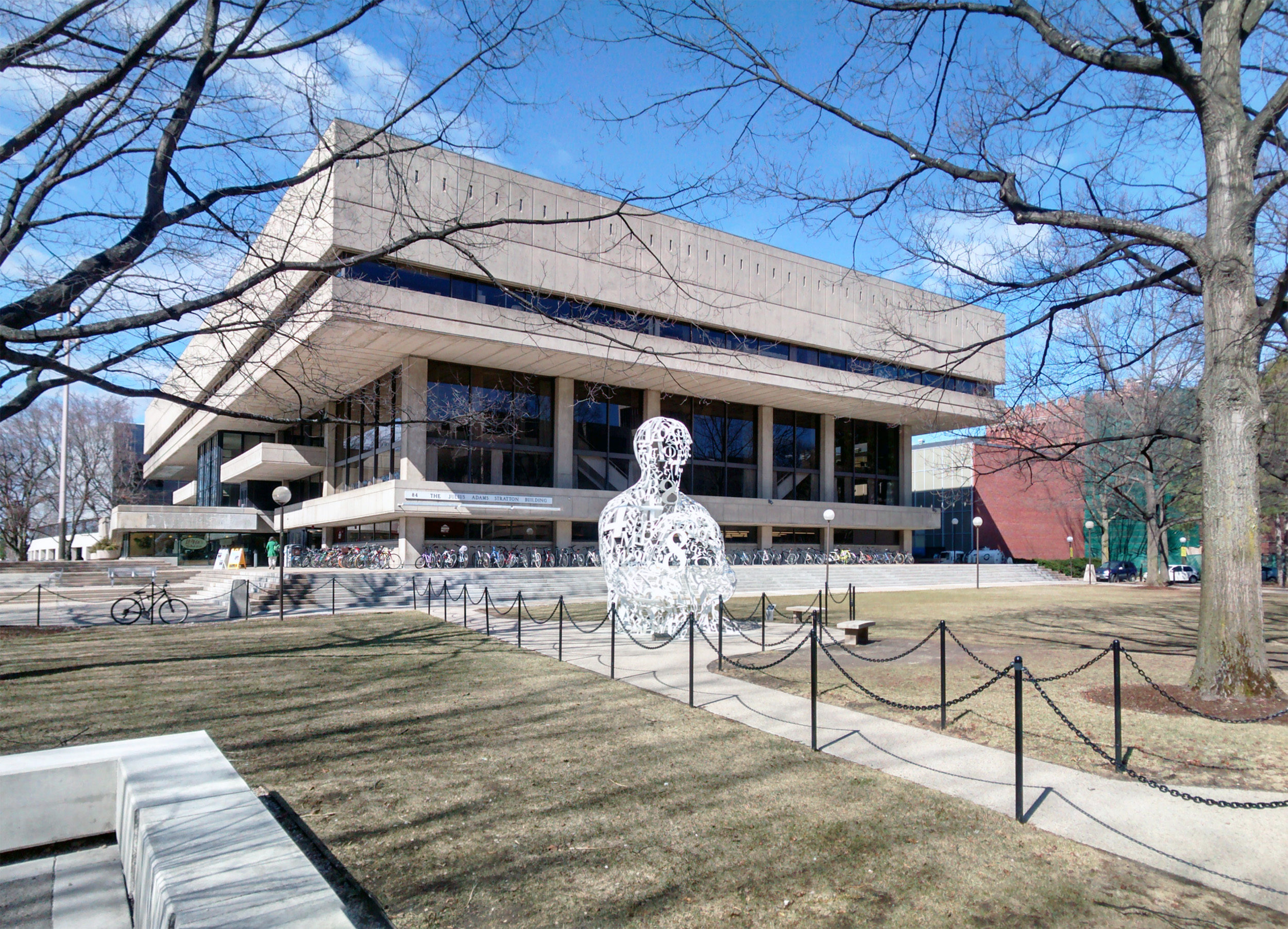Grey building with the top floor jetting out above balconies on all sides. The alchemist sculpture is in front of the building on a green lawn.
