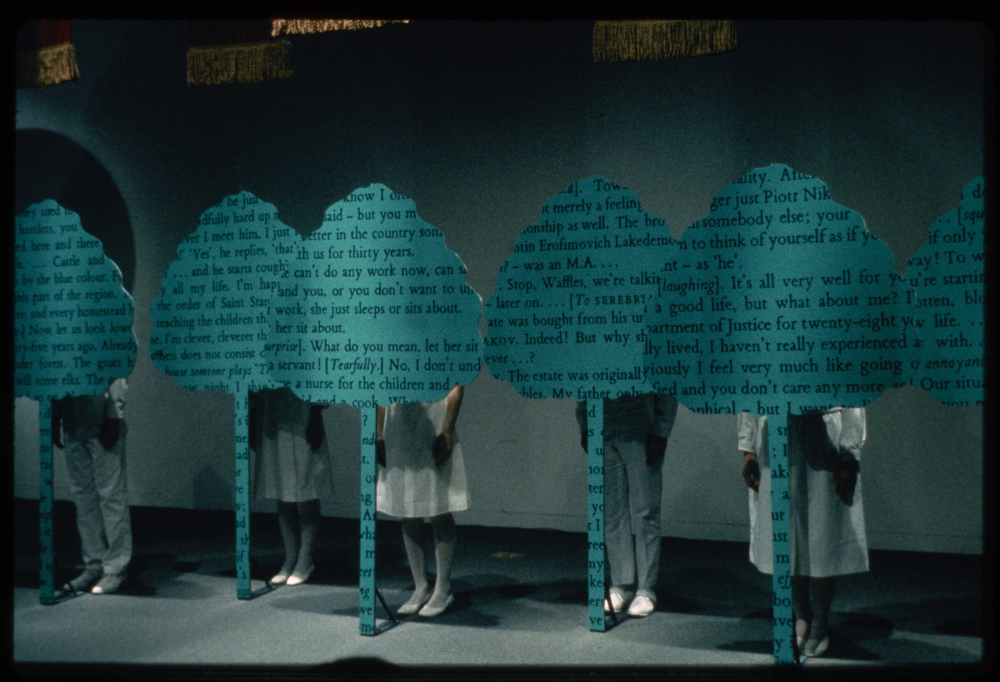 Performers dressed in white stand behind cout-outs of trees covered in text within the installation.